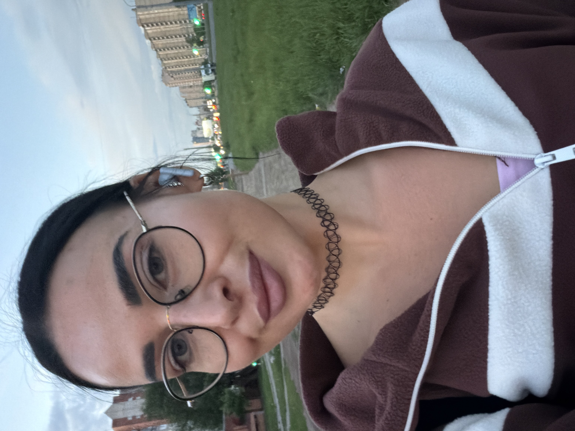 An outdoor selfie taken by a person, presumably a woman, in what appears to be Saint Petersburg, Russia. The image is oriented sideways, but depicts the subject looking directly into the camera with a neutral to slightly pleasant expression. The woman has dark hair pulled back and wears round, black-framed glasses, framing prominent dark eyebrows. A black, woven choker-style necklace is visible around her neck. She is dressed in a brown fleece-like jacket with a thick white stripe running down the front, which is partially unzipped to reveal a light purple undergarment beneath.
The background indicates an urban setting at what appears to be dusk or during an overcast day. Above the woman's head (if the image were upright), a light, cloudy sky is visible, with several multi-story buildings forming a distant cityscape. Below and behind her (if upright), a stretch of vibrant green grass runs alongside a paved path or sidewalk. Further in the background, more urban structures line the horizon, interspersed with blurred artificial lights glowing green and reddish-orange, suggesting streetlights or traffic signals. The overall atmosphere is calm, captured during an outdoor moment in an urban green space. There is no visible text in the image.