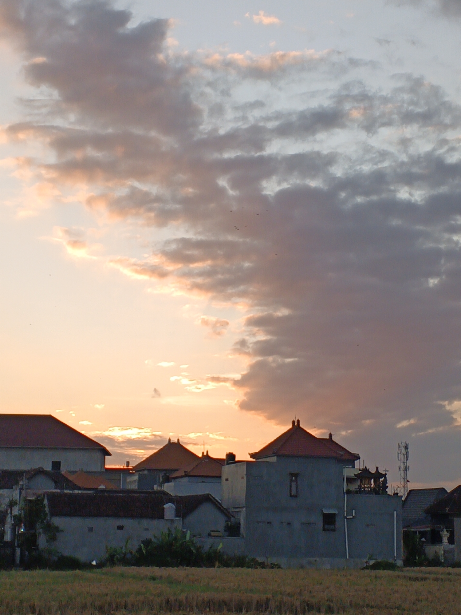 The image depicts a rural landscape in Sukawati, Indonesia, at dusk. The sky is filled with a mix of soft orange and pink hues from the setting sun, overlaid with dramatic, dark gray clouds. In the foreground, dry, straw-like vegetation covers the ground, suggesting a harvested field. A cluster of buildings with reddish-brown tiled roofs and gray concrete walls forms the middle ground. One building, taller and more modern, features small rectangular windows and a cell tower on its roof. The structures appear to be residential homes or small businesses. The overall scene conveys a sense of quiet and the end of the day in a tropical setting.