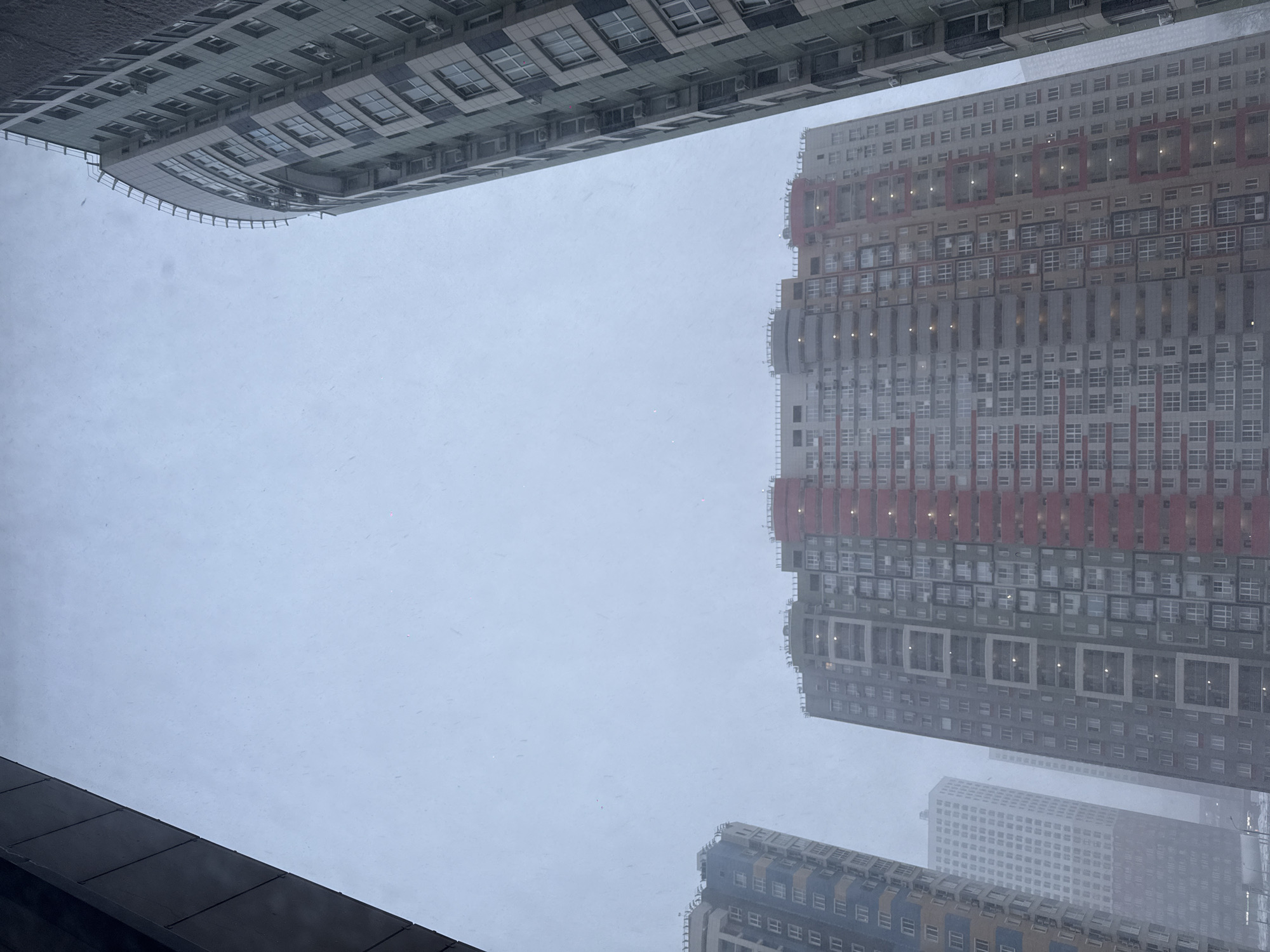 The image depicts a view upwards towards tall buildings in what appears to be a city. The sky is overcast and it seems to be snowing. The buildings have multiple stories with numerous windows. One building has sections of red detailing. The location is Khimki, Russia.