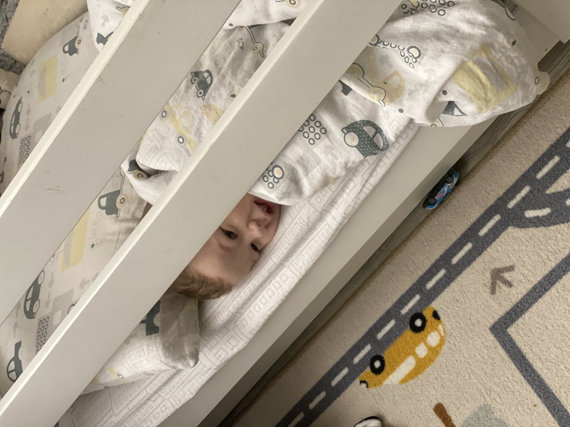 A young child lies in a crib, partially covered by a blanket with a car-themed pattern. The crib has white slats. A blue toy car is resting on the side of the crib, near a rug with a road pattern. The child's face is visible between the slats. The location is not discernible from the image.