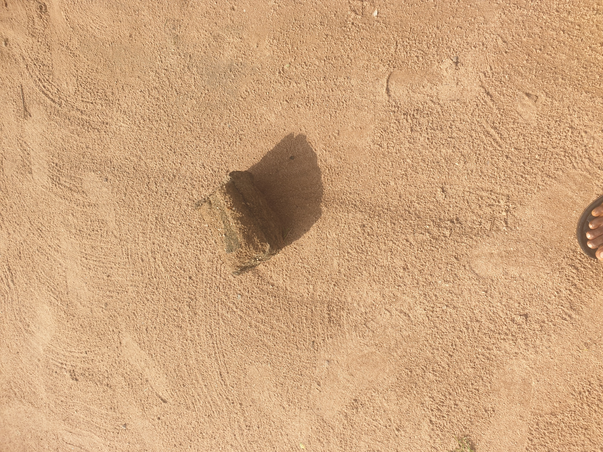 The image captures a close-up, high-angle view of a sandy area, likely a beach or outdoor ground, in Jalingo, Nigeria. The sand is a light brown or tan color and shows signs of having been walked on, with several faint footprints visible. In the center of the frame, a rectangular piece of what appears to be a brick or a piece of concrete rests on the sand, casting a shadow to its right. The texture of the sand is granular and uneven, with some areas showing ripples or patterns from wind or water. The lighting suggests it is daytime, with the sun casting distinct shadows. At the bottom edge of the image, a portion of a bare foot wearing a sandal is visible, indicating the presence of a person. The overall scene is simple and natural, focusing on the texture of the sand and the solitary object.
