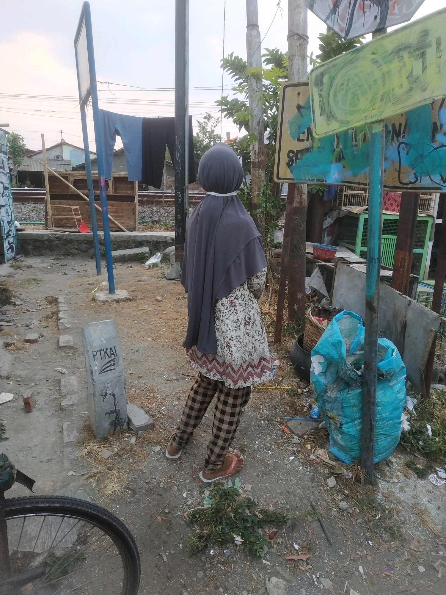 A person wearing a dark grey hijab, a patterned long-sleeved shirt with a white zig-zag trim at the bottom, and checkered brown and black pants stands on a dirt path near a railway track. The person is facing away from the camera, towards a blue plastic bag filled with debris, a woven basket, and a red bucket. In the background, laundry hangs on a clothesline between two poles, and a concrete marker with the inscription "PTKAI" is visible. The environment appears somewhat disheveled, with scattered debris and dry vegetation. The scene is likely in Tegal, Indonesia, given the "PTKAI" marking, which is associated with Indonesian state-owned railway company PT Kereta Api Indonesia.