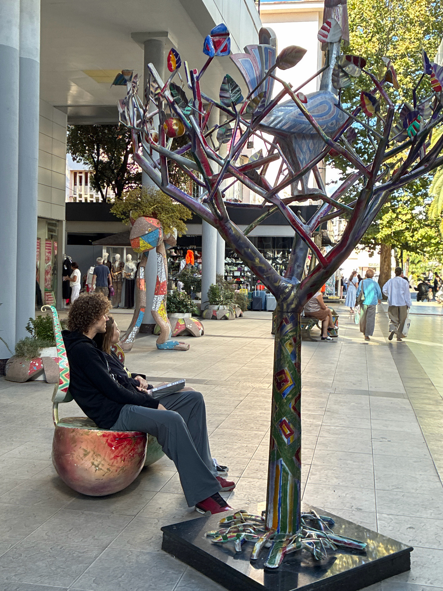 The image depicts an outdoor pedestrian area, likely a promenade or shopping street. In the foreground, a colorful, stylized tree sculpture with abstract leaves and a bird-like element dominates the view. The sculpture is mounted on a black base. To the left, a couple is seated on a unique, round bench shaped like a stylized apple. The man has curly hair and is wearing a black hoodie and gray pants, while the woman, partially visible, is wearing a yellow top and jeans. A laptop rests on the woman's lap.
In the background, a large, mosaic-covered sculpture of a humanoid figure stands. Various shops with mannequins are visible behind the sculptures. Several people are walking in the distance, including two women in light-colored clothing and a man in a light shirt. The ground is paved with square tiles. The environment appears to be a well-maintained urban public space, suggesting it is a tourist or commercial district. The architecture in the background includes a building with white columns and light-colored facades, indicative of a modern construction. The presence of trees and sunlight suggests a pleasant weather day. The location is identified as the Arbat street in the city of Sochi, Russia.