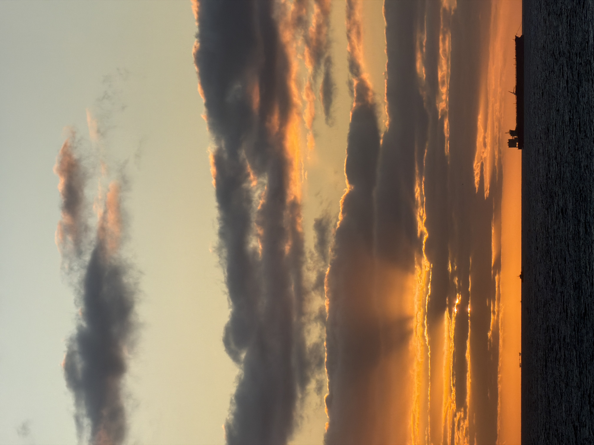The image depicts a sunset scene, likely over water, in an unknown city and location. The sky is filled with dramatic clouds, some dark grey and others illuminated with orange light from the setting sun. There is a visible horizon line where the sky meets the water. A silhouette of what appears to be a large ship or structure is present on the horizon. The water itself is dark and textured. The overall color palette is dominated by shades of orange, grey, and dark blue.