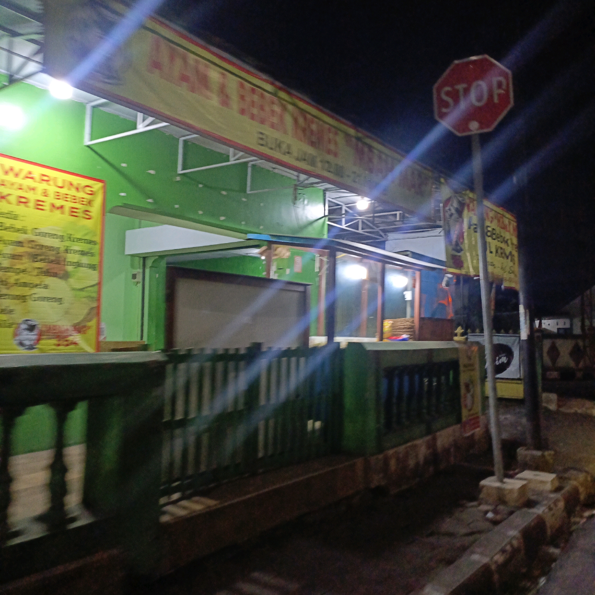 This is a night-time outdoor image of a street scene in Salatiga, Indonesia. The primary focus is a food stall with a prominent yellow banner that reads "AYAM & BEBEK KREMES" and "BUKA JAM 10.00-21.00". This translates to "Chicken & Duck Kremes" and "Open 10:00 AM - 9:00 PM". The stall is illuminated by artificial lights, creating a bright green backdrop. To the right of the stall, a red octagonal stop sign is visible. A wooden fence runs across the foreground, separating the viewer from the stall area. The ground appears to be paved, with some debris visible. The overall impression is that of a casual dining establishment operating in a local urban setting.