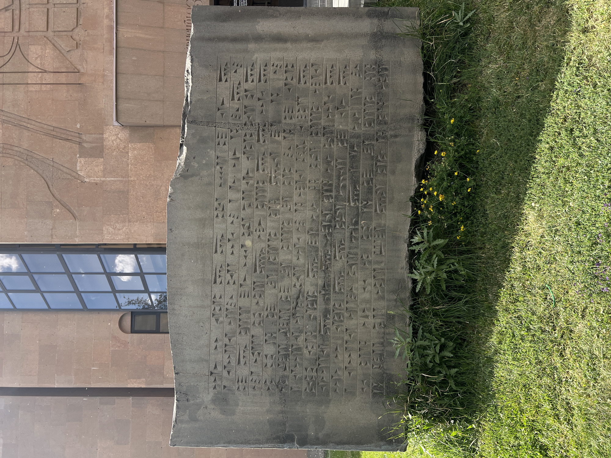 The image displays a large, dark grey stone slab or stele, prominently featuring deeply carved cuneiform inscriptions. The ancient script is meticulously arranged in numerous vertical columns, delineated by both horizontal and vertical lines, with individual wedge-shaped characters clearly visible across the weathered surface of the stone.
The slab is positioned horizontally on the ground. To its left, a modern building constructed from light-colored, possibly tuff, stone rises. This building's facade includes geometric carved patterns and a large, multi-paned window that reflects a bright, partly cloudy sky, indicating a sunny day. To the right of the slab, a strip of vibrant green grass grows, interspersed with wild plants and small yellow flowers.
No people are visible in the scene. The monumental, historical artifact with its ancient text stands in stark contrast to the contemporary architectural style of the adjacent building. The visible text on the slab is the cuneiform script itself. The location context is Yerevan, Armenia.
