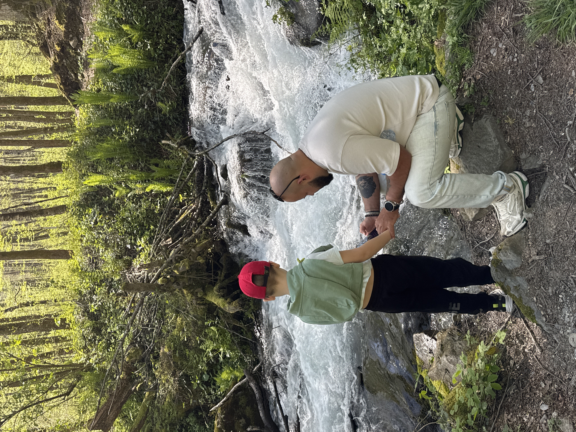 The image depicts a man and a child near a waterfall. The man is bald, wearing a light-colored shirt and light-colored jeans, and is kneeling with one knee on a rock. He has a tattoo on his arm and a watch on his wrist. The child is wearing a red cap, a light green jacket, and black pants. They are holding hands near the waterfall. The environment consists of a fast-flowing waterfall surrounded by lush greenery, rocks, and trees. The location is likely in a natural area near Krasnaya Polyana, Russia, given the vegetation and terrain.