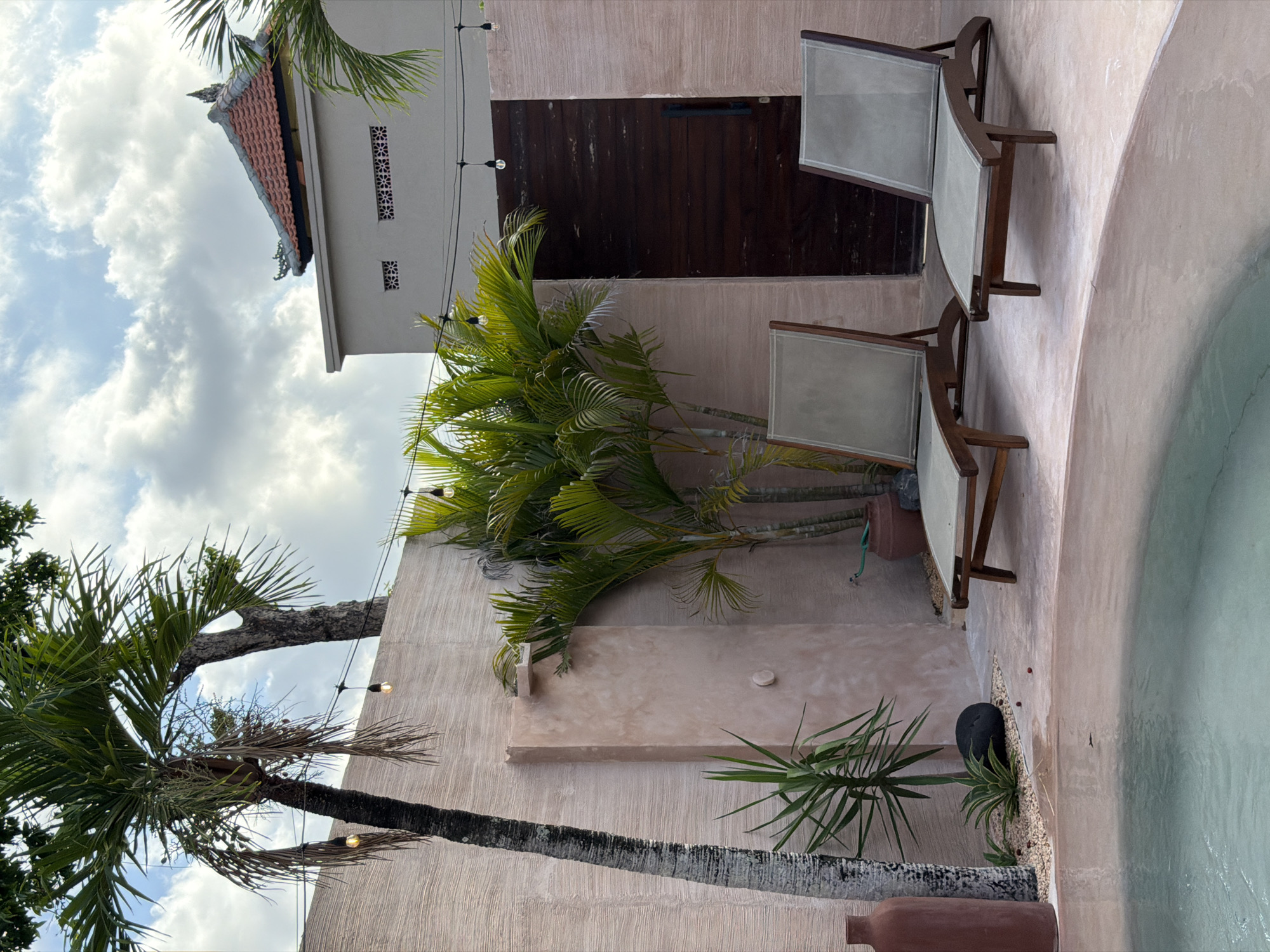 The image depicts an outdoor scene, likely a private residence, located in North Kuta, Indonesia. The composition is dominated by a light pink wall constructed with a rough, textured stucco. Palm trees are prominent, one large tree is situated on the left, partially obstructing the view.
A pair of wooden framed lounge chairs with light-colored canvas are placed against the wall. Next to the chairs is a partially visible pool with bright blue water.
String lights are strung across the building. A section of the building has a wood-paneled section, possibly a gate or large door. The sky is filled with puffy white clouds. A small architectural building is visible in the background.