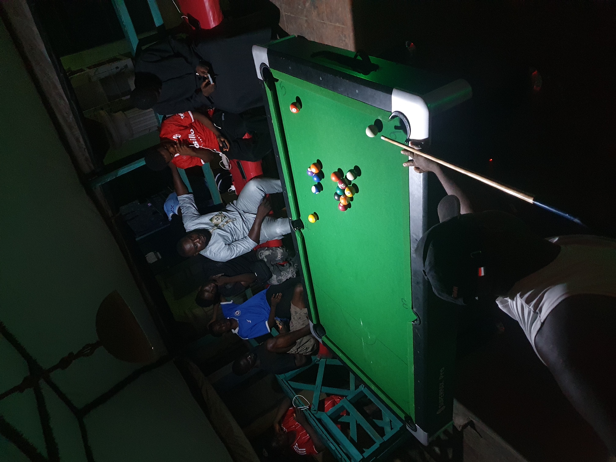The image captures an indoor scene in Lokoja, Nigeria, featuring a group of individuals gathered around a billiards table. The pool table, covered in green felt, is the central object, with a set of pool balls clustered towards the middle. A person is in the act of taking a shot, cue stick in hand, aiming at the cue ball. Other individuals are lounging around the table, some sitting on chairs or the floor, observing the game. The environment is dimly lit, and the space appears to be a recreational area, possibly a bar or a private game room. Some of the individuals are wearing casual attire.