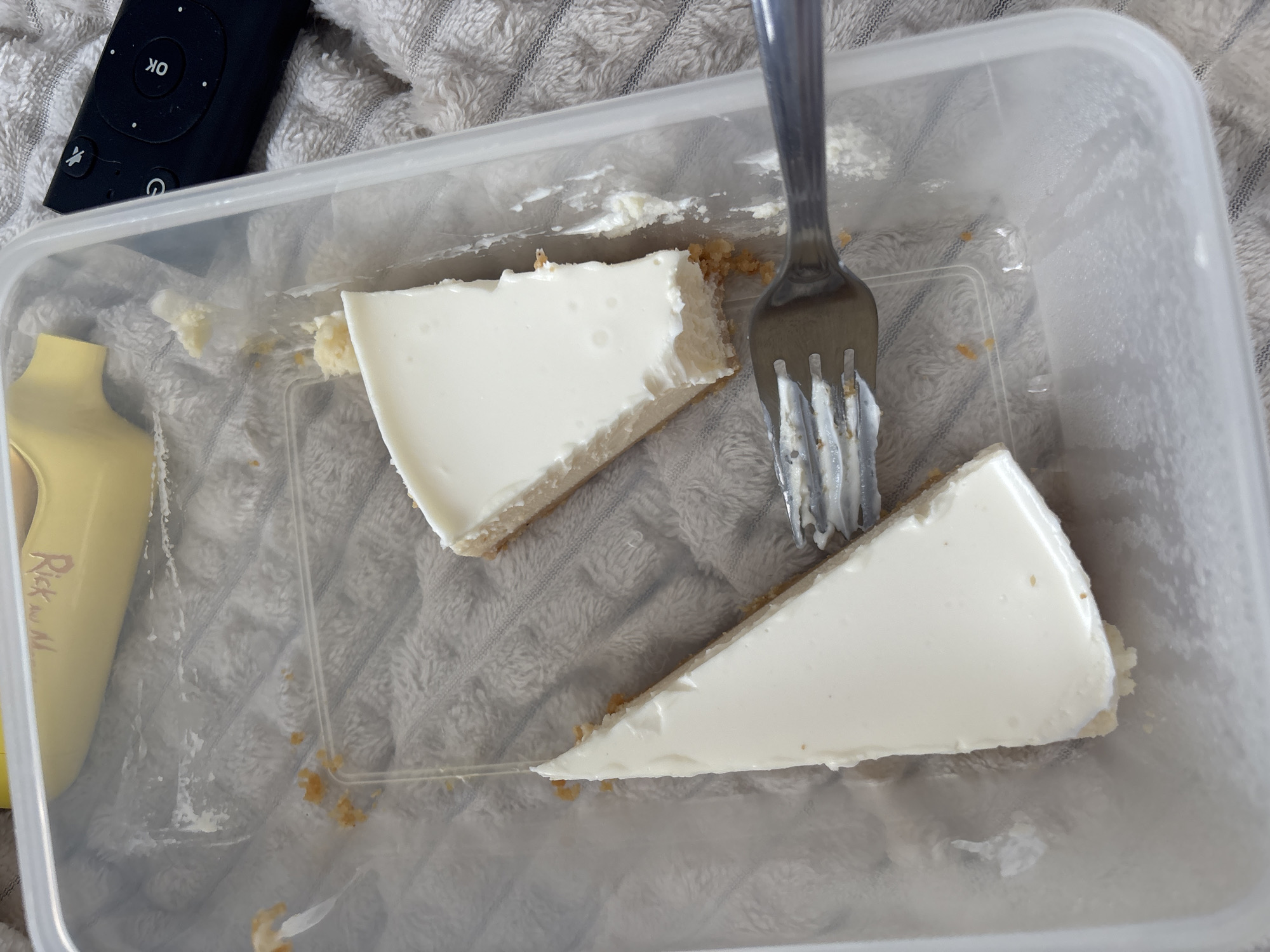 The scene presents a partially consumed cheesecake in a clear plastic container, accompanied by a fork with cheesecake residue. The container rests on a textured, light-colored surface. A black remote control lies adjacent to the container. The details of the image do not indicate any location in the city of Vsevolozhsk, Russia. There is no forensic information available in the image, and any description of this image should be considered observational.