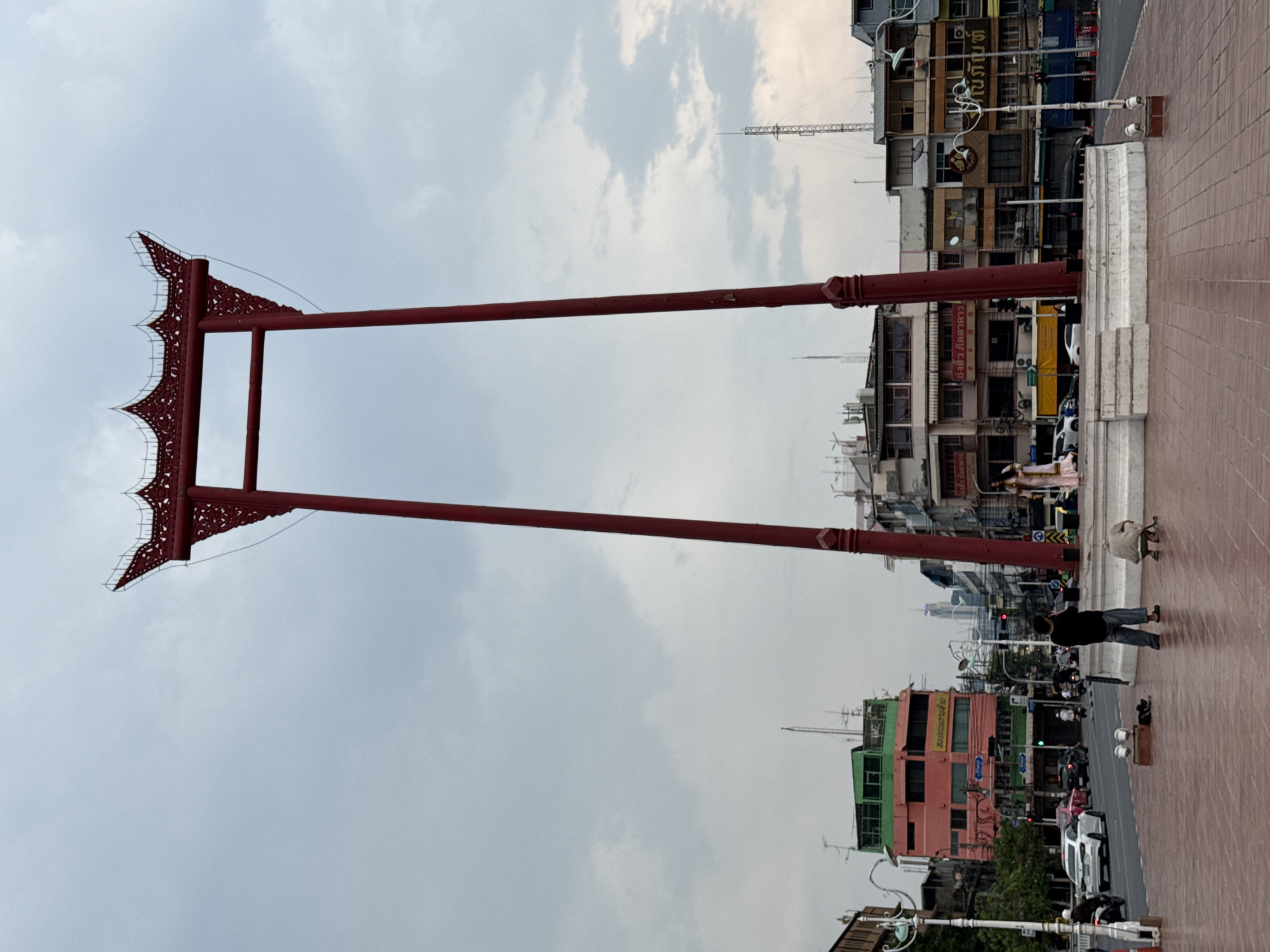The image shows the Giant Swing, a religious structure in Bangkok, Thailand. It consists of two tall, red painted pillars with an ornate, decorative frame at the top. The structure stands on a brick-paved ground. Buildings of varying heights and colors surround the structure. There are people walking on the ground. The sky is cloudy.