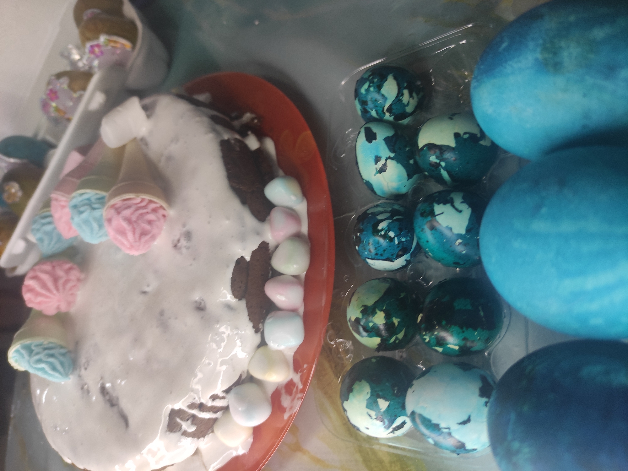 The image shows a table with Easter decorations. A chocolate cake with white icing and pastel-colored marshmallow decorations sits on a red plate. To the right of the cake are several blue-dyed Easter eggs in a clear plastic carton. There is another container on the left side with pastel candy ice cream cones in it. The picture seems to have been taken indoors. The location is not determinable with the given information.
