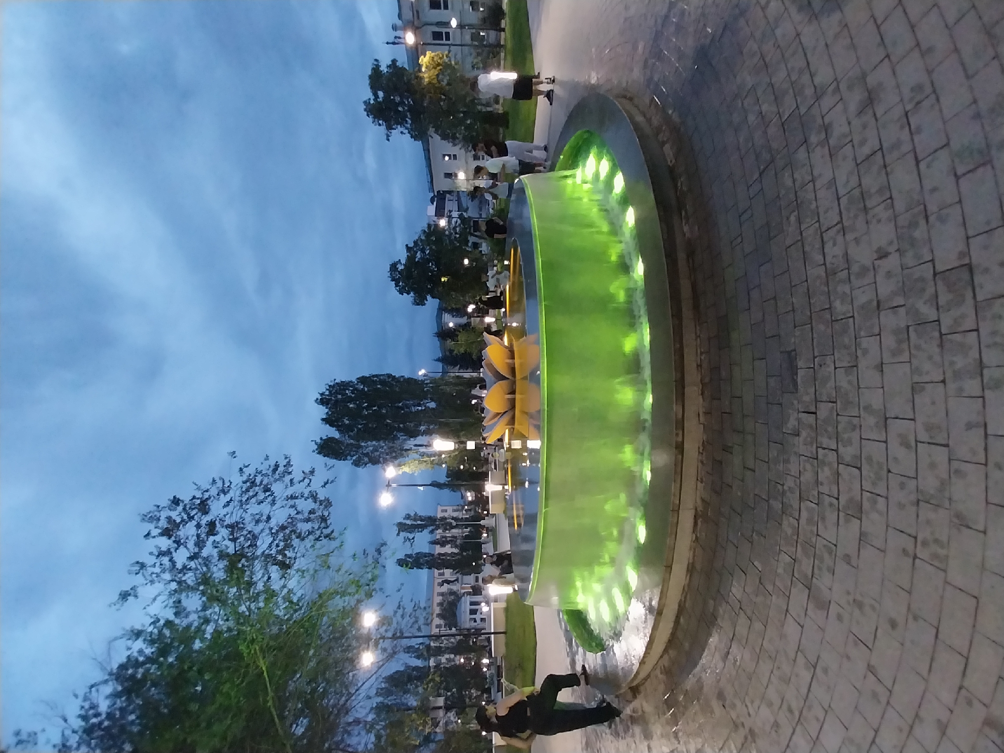 This image depicts an outdoor fountain at dusk, with green water illuminated by underwater lights. The fountain's rim is made of stone, and the surrounding area is paved with interlocking grey bricks. Several individuals are present, some standing near the fountain and others in the background, which includes trees, lampposts, and buildings. The sky is overcast and a deep blue, suggesting the twilight hour. The fountain's design features a central element resembling a lotus flower. Based on the provided context, this scene is located in the city of Orenburg, Russia.