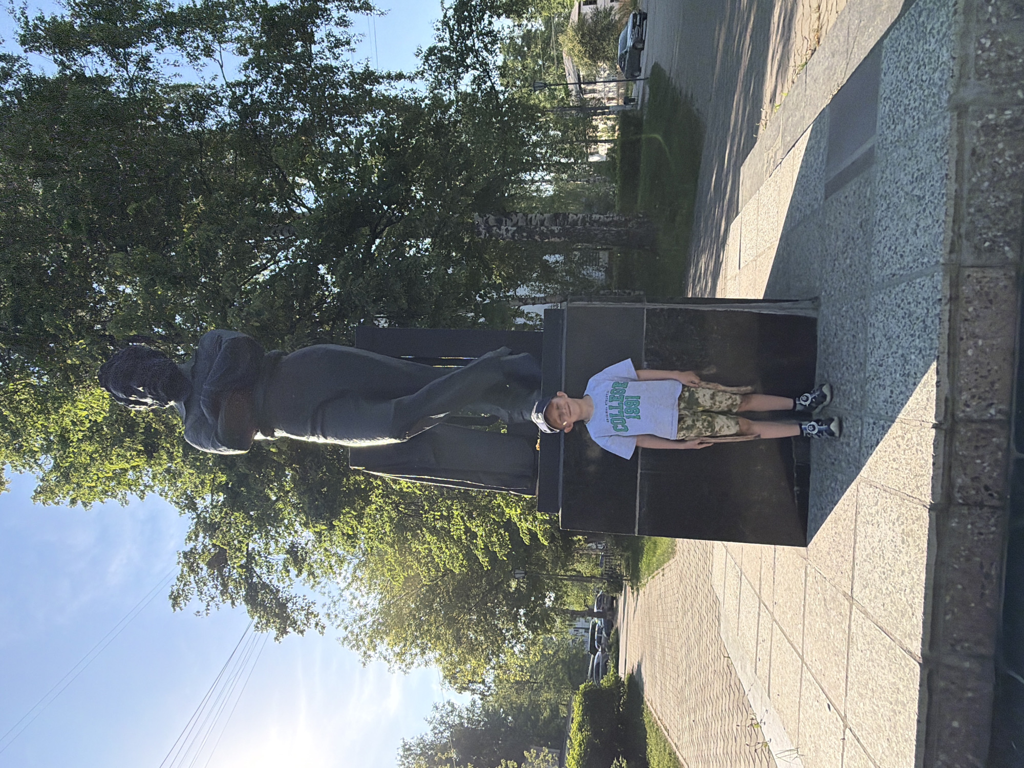 A child wearing camouflage shorts, a white t-shirt, and a white baseball cap is standing on the base of a bronze statue of a man. The statue stands in a city park with trees in the background. The location is Chaikovsky, Russia.