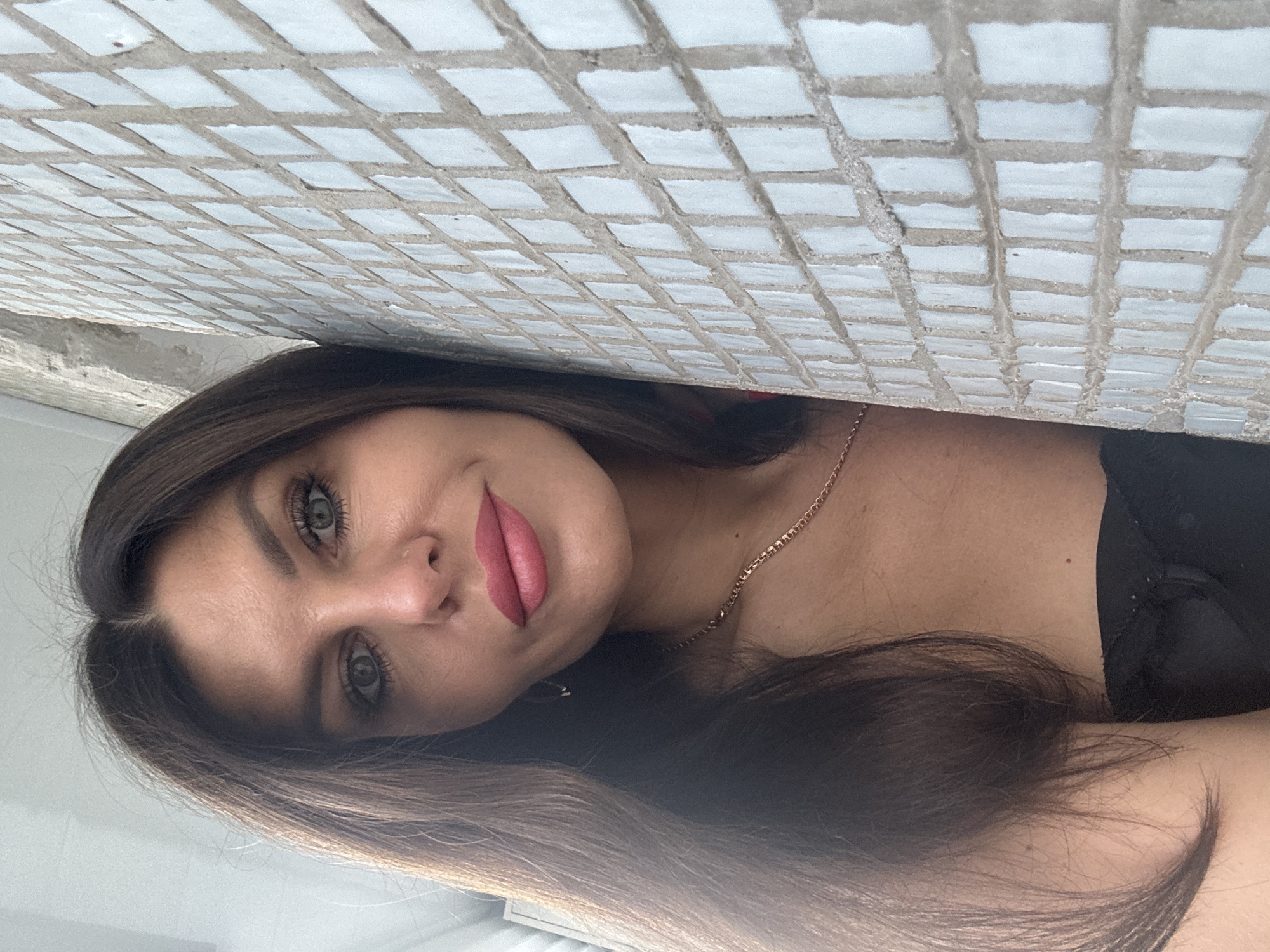 A close-up, outdoor, eye-level, natural light selfie shows a woman with dark brown hair and fair skin. She is looking directly at the camera, tilting her head to her left. Her hair is long and wavy, falling over her left shoulder. She is wearing pink lipstick and has green eyes. A thin gold necklace is visible around her neck. She is wearing a black top. The background consists of a light-colored tiled wall to her right and a rough, light-colored concrete structure to her left. The image was taken outside, with the sunlight casting shadows on the wall. The woman appears to be in a public space, possibly an alleyway or a courtyard, given the wall and concrete structures.