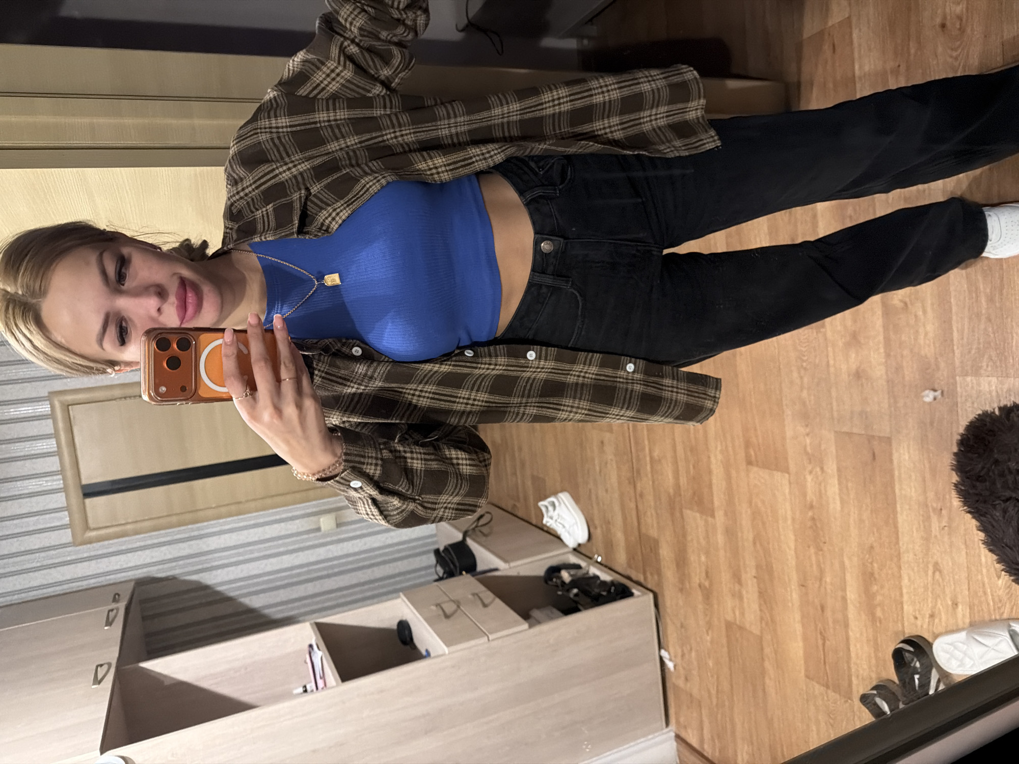 A young woman with blonde hair pulled back takes a selfie in a room. She is wearing a blue crop top, a brown plaid open shirt, and black jeans. A gold necklace with a rectangular pendant is visible around her neck. She is holding a smartphone in her right hand, which is taking the picture. The room appears to be a bedroom or dressing area, with a closet, shelves, and a door visible in the background. The floor is made of light-colored wood. There are shoes on the floor near the closet. The overall impression is casual and domestic.
