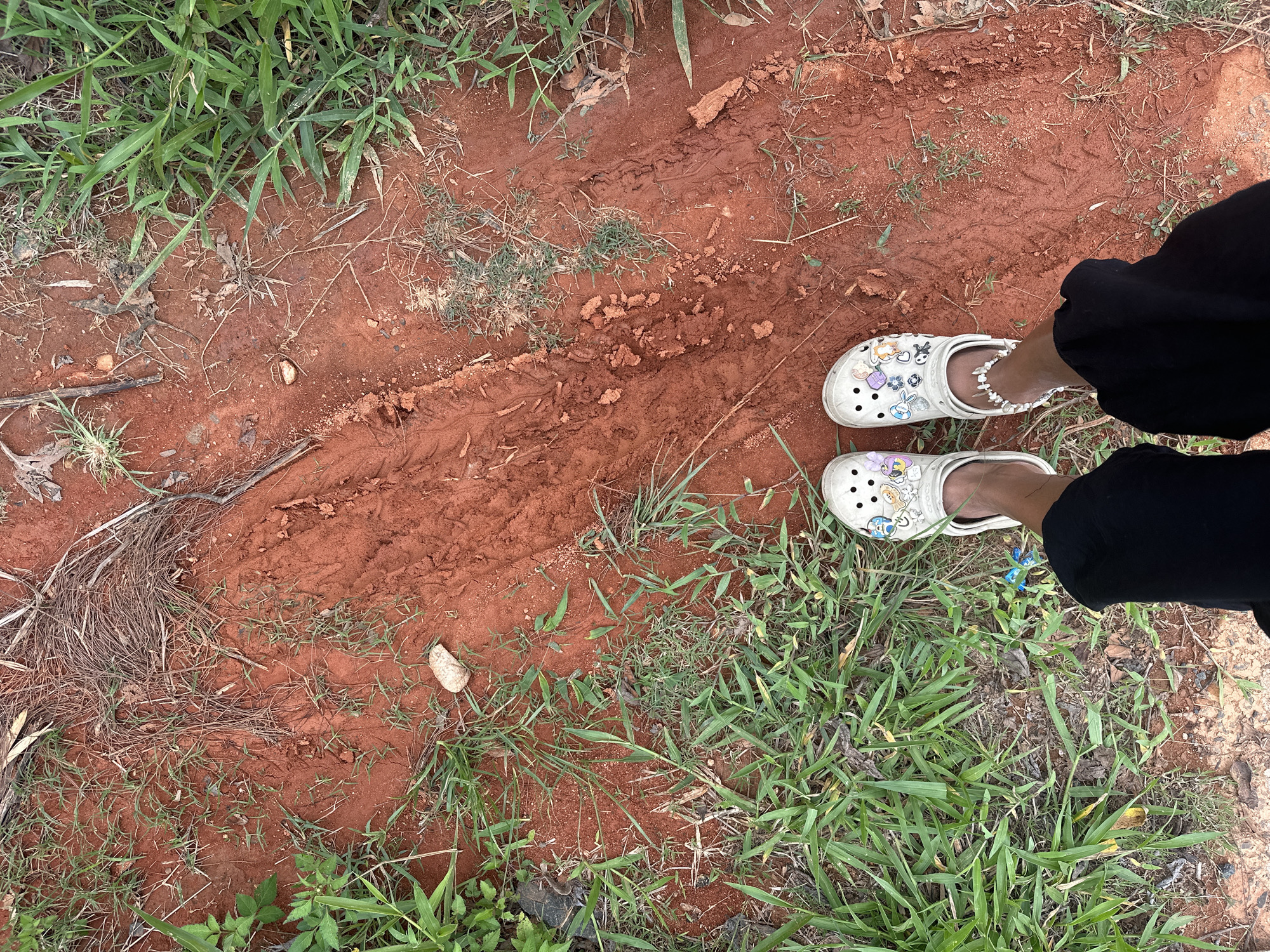 A ground-level view of a patch of red soil in Da Lat, Vietnam. The soil is uneven, with tire tracks visible. Patches of grass grow along the edges of the soil. Two feet clad in white Crocs sandals with decorative charms stand on the soil. The individual is wearing black pants.