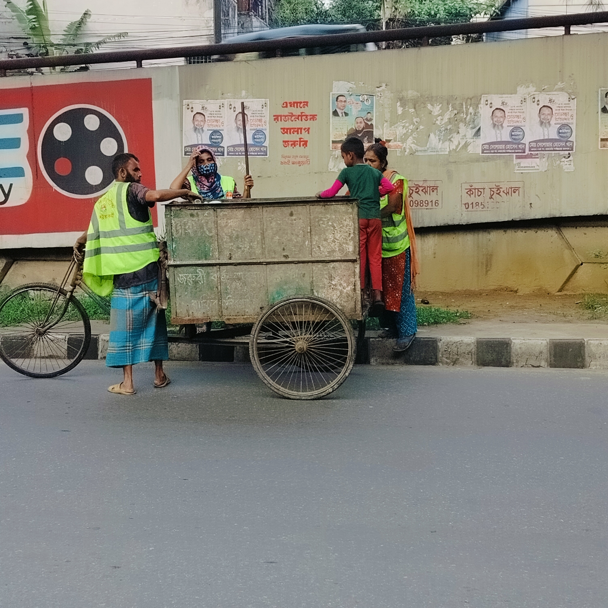 The image shows a street scene in Dhaka, Bangladesh. Three adults and one child are gathered around a large, wheeled metal cart. A man stands next to the cart holding a bicycle. He is wearing a safety vest over a dark shirt and a blue and white checkered lungi, with sandals on his feet. A woman inside the cart holds a stick. She wears a blue floral face covering and a safety vest. A second woman, also in a safety vest, stands next to the cart, seemingly assisting a young boy in red pants and a green shirt who is climbing into the cart.
The cart is positioned on the edge of a paved road, alongside a concrete curb. The background consists of a wall covered with posters and advertisements in Bengali. Several political posters are visible. A large red and white sign with a black circle design is visible on the left side. Trees are visible above the wall. The road surface is cracked. The photograph appears to have been taken during the daytime, based on the lighting. A sign on the wall reads "Kacha Chuihal 01851".