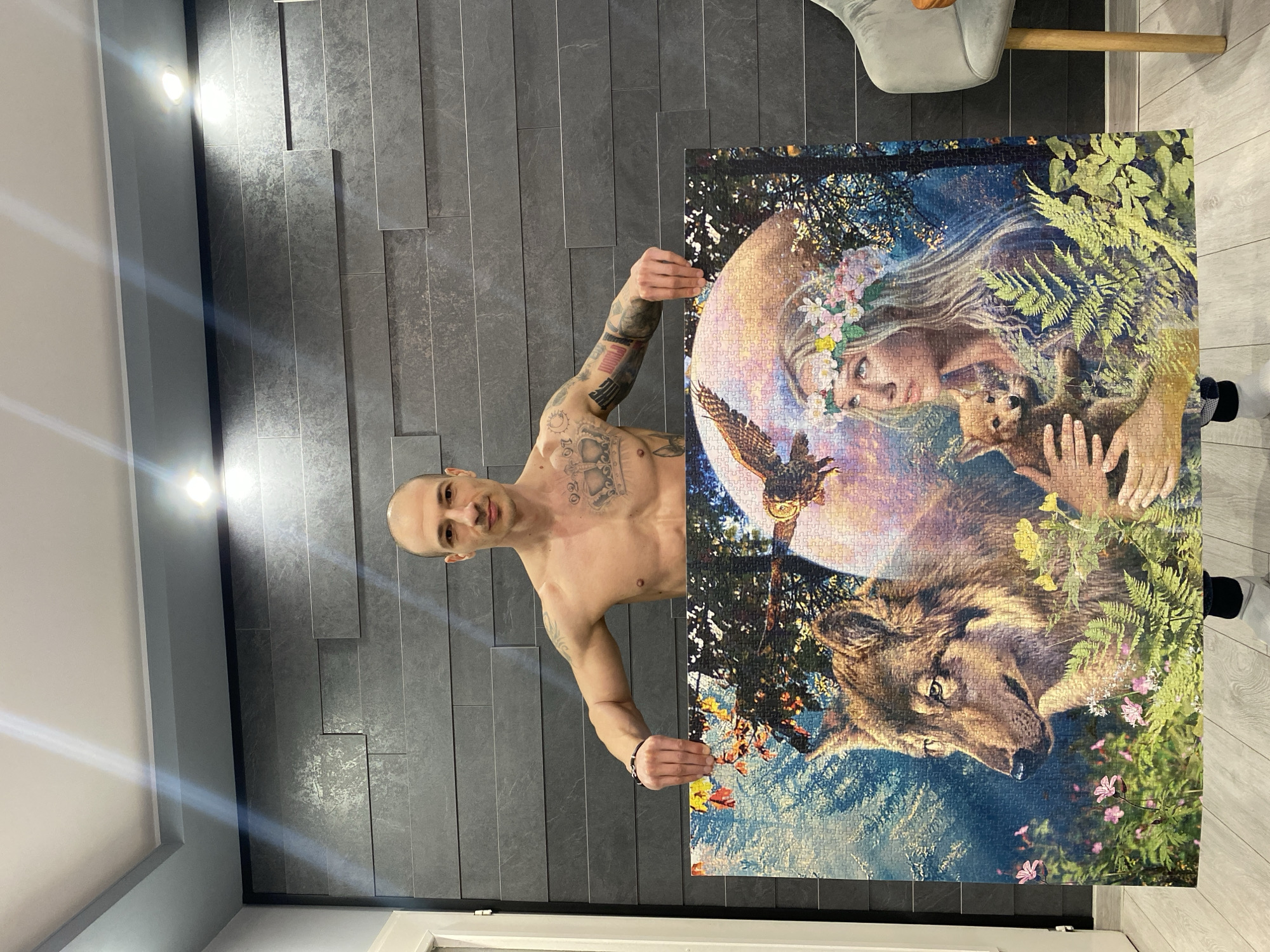 A man with a shaved head and tattoos is holding up a large jigsaw puzzle in front of a grey textured wall. He is shirtless and facing the camera. The puzzle depicts a woman with a flower crown, a fox, an owl, and a wolf. The location appears to be indoors, with a chair visible in the background. The scene is lit by overhead lights. The man is standing in what appears to be a living space. There is no indication of the city of Pleven, Bulgaria in the image.