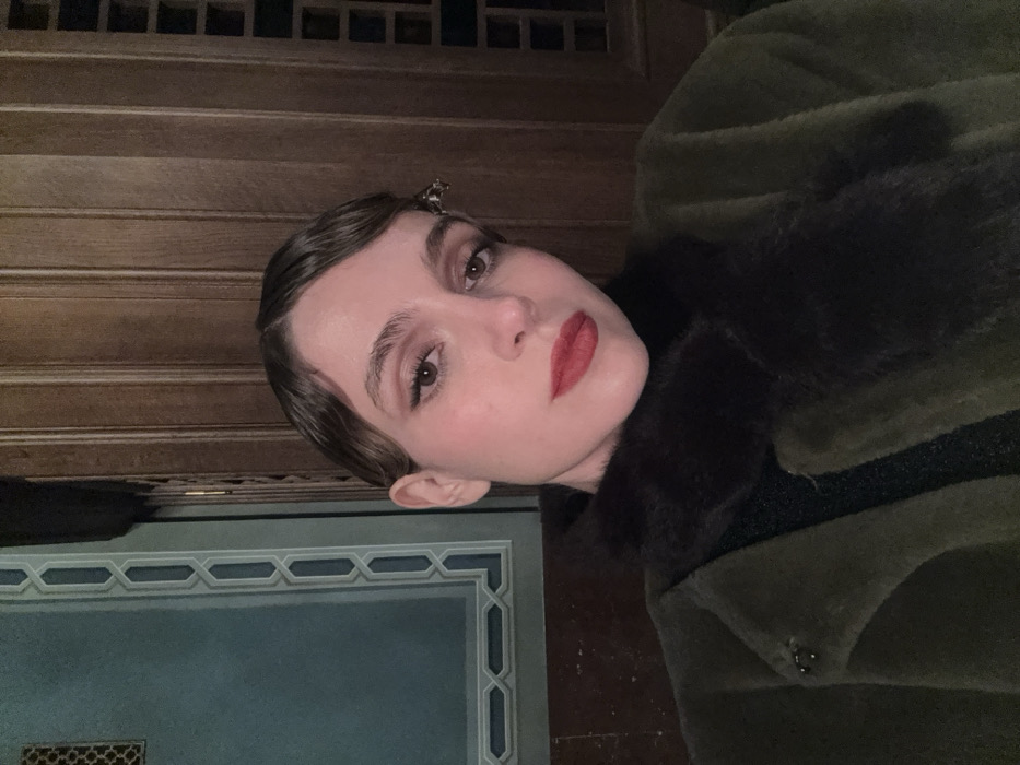 A woman poses, wearing a green fur coat with a dark fur collar, red lipstick, and a hair clip. Her hair is styled in vintage waves. The background consists of a dark wooden wall and blue wall with a decorative border. The picture is angled, giving a tilted perspective.