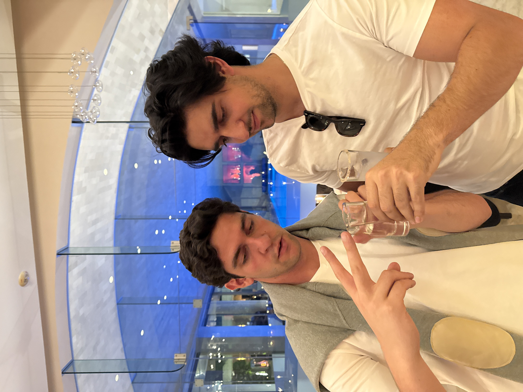 Two young men are standing in what appears to be a modern shopping mall. The man on the right is wearing a white t-shirt and has a pair of sunglasses hooked on his shirt. He is holding a glass, as if pouring liquid into another glass being held by the other man. The man on the left is wearing a light grey sweater over a white shirt, and is holding his hand up with two fingers extended in a "peace" sign. They are both looking down, possibly at the glasses. The background consists of multiple floors with reflective glass, white paneling, and blue lighting accents, creating a sleek and modern aesthetic. There is a decorative hanging light fixture visible in the upper left of the frame. The reflections in the glass hint at shops and activity in the mall. Based on the style of architecture and lighting, it is likely this photo was taken at a modern shopping mall in Mexico City, Mexico.