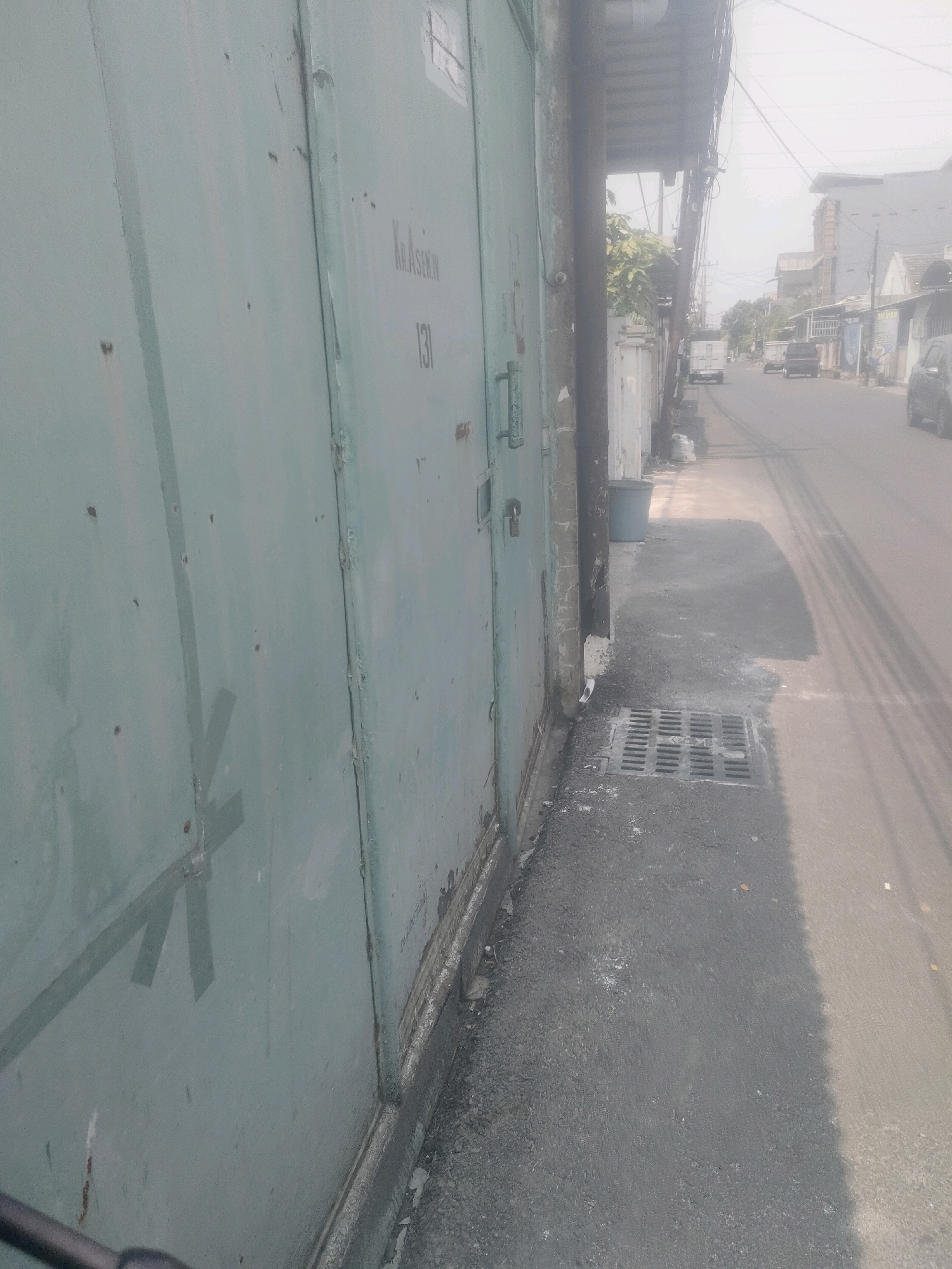 The image shows a street scene in Surabaya, Indonesia. On the left, a large, weathered, pale green metal gate occupies the foreground, exhibiting signs of wear and tear, including rust and several indentations. The gate has visible seams, hinges, and what appears to be a lock mechanism. Faint lettering and the number "131" are visible on the gate. To the right of the gate, a concrete sidewalk runs alongside the street, bordered by a drain grate. A grey plastic trash bin is situated on the sidewalk. The street itself is paved with asphalt and shows tire marks. A utility pole with numerous wires stands near the gate. In the background, several buildings and vehicles are visible, including white vans and cars, suggesting a typical urban environment. The overall lighting indicates it is daytime, with the sky appearing hazy.