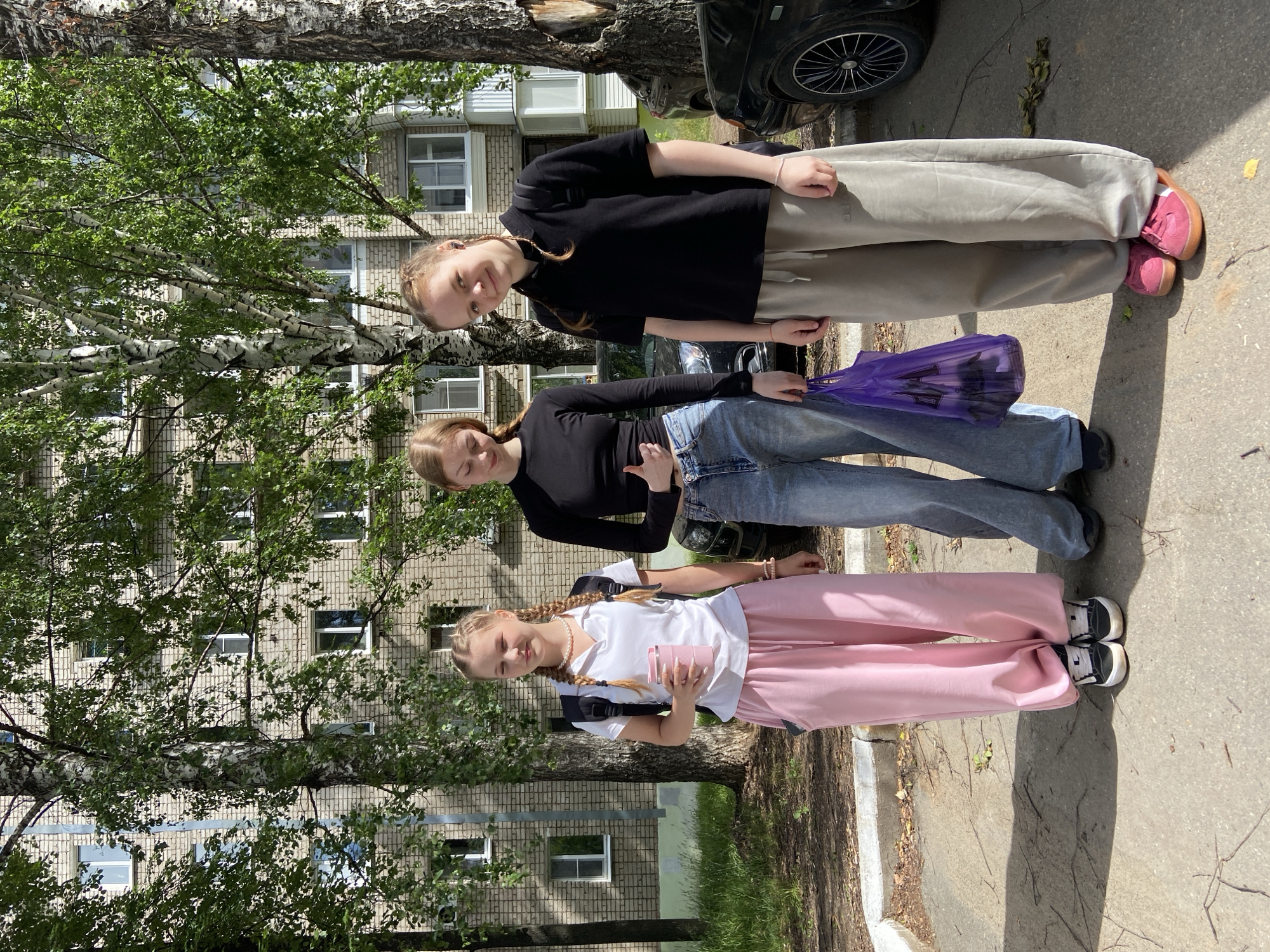 Three young females are standing in front of a multi-story brick building with trees and parked cars. The female on the left is wearing a white polo shirt, pink sweatpants, and black and white sneakers, and is holding a pink cup. The female in the middle is wearing a black long-sleeved shirt, light blue wide-leg jeans, and dark sneakers, and is carrying a purple plastic bag. The female on the right is wearing a black polo shirt, light grey sweatpants, and pink sneakers, and has a backpack. The scene appears to be in an outdoor urban environment. The architecture of the building suggests it may be a residential building in Chaikovsky, Russia, likely from the Soviet era or later.