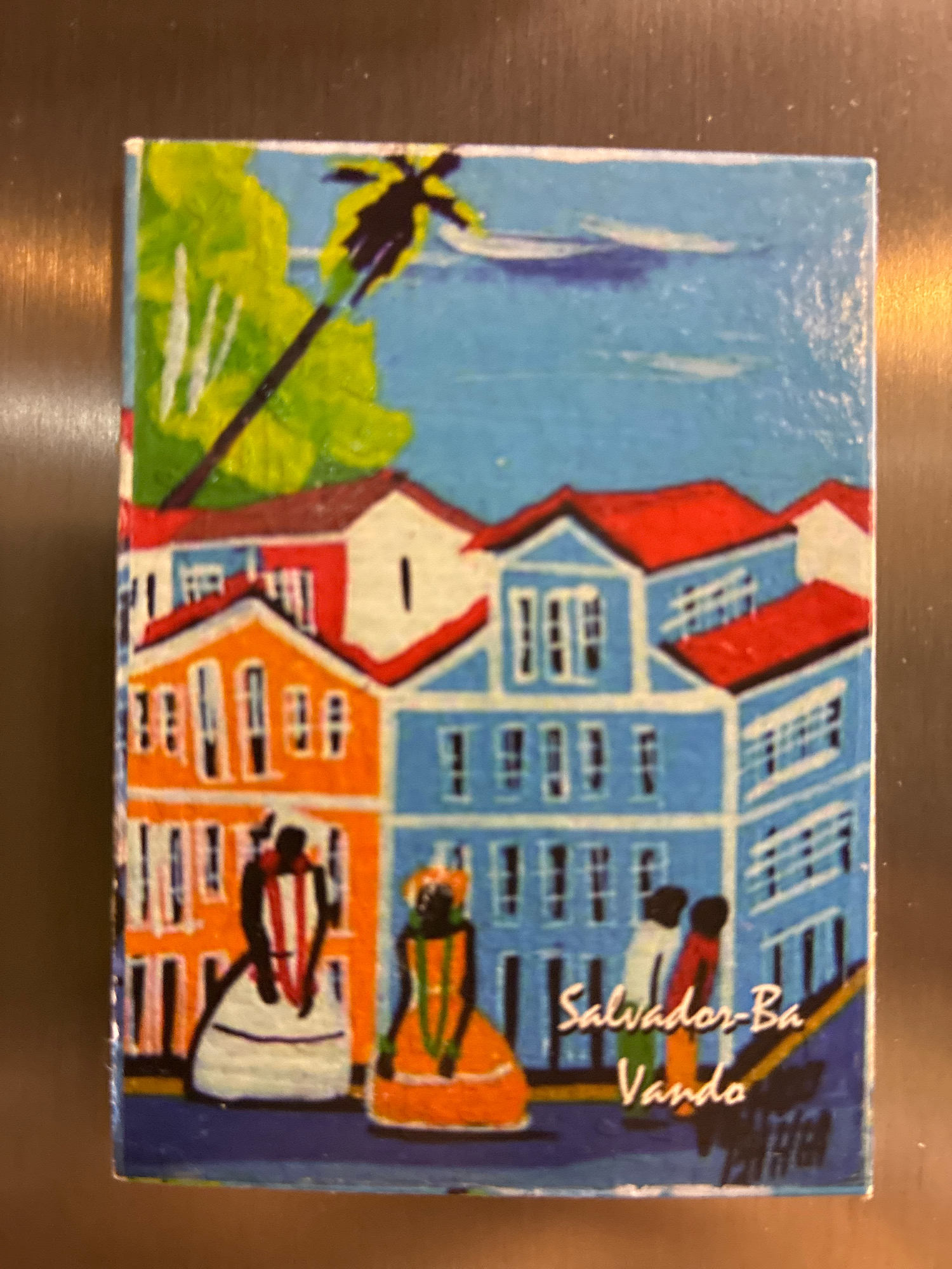 The image is a painting of colorful buildings in a city. The buildings are painted in shades of blue, orange, and white, with red roofs. There are some figures painted in the foreground, including two people in traditional clothing. The painting also features a palm tree and a bright blue sky. The text on the painting reads "Salvador-Ba Vanda". The painting appears to be a souvenir magnet. This painting does not depict any specific location in the city of Curitiba, Brazil.
