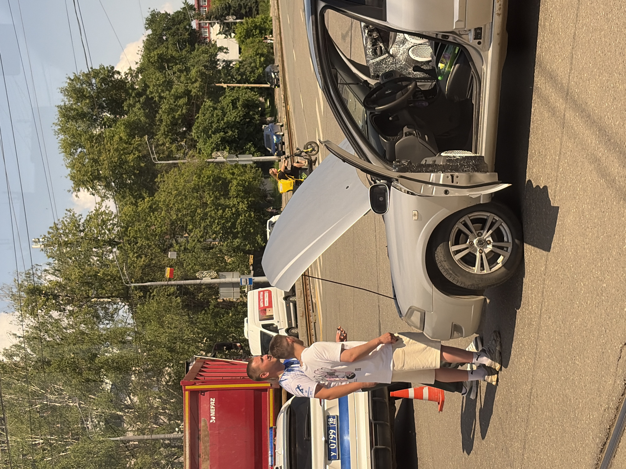 The image depicts a street scene in Izhevsk, Russia, likely during daytime. In the foreground, a silver car with its hood open is parked. Two men are standing near the car, one in a white t-shirt and light-colored shorts, and the other in a blue and white t-shirt and dark pants. A white police car is parked to the left of the silver car, with its emergency lights activated. A red truck with "NEFAZ" branding is visible behind the police car. In the background, trees and buildings line the street, and traffic signals are present. A person on a scooter with a yellow backpack is also visible in the mid-ground. The overall scene suggests a traffic incident or breakdown situation.