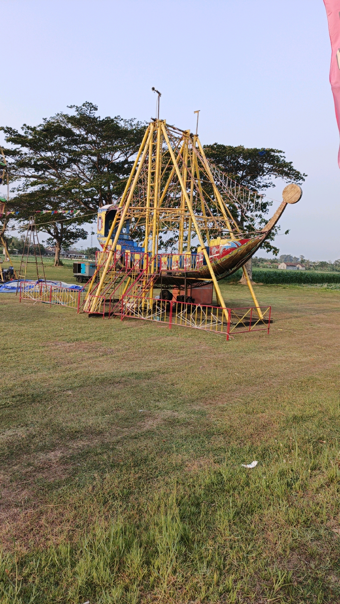 An amusement park ride, designed to resemble a pirate ship, stands on a grassy field under a clear sky. The ride's main structure is a bright yellow metal frame supporting a large, boat-shaped car that hangs from a central pivot. The boat is painted with colorful designs, including red, blue, and yellow patterns. Red and yellow metal barriers surround the base of the ride, creating a queueing area. Several steps lead up to the entrance of the boat.
In the background, large trees with dense foliage are visible, along with a few buildings and a green field that appears to be cultivated with crops, possibly corn. The overall environment suggests an outdoor recreational area. There are no people actively engaged in riding the amusement park attraction in this moment. The scene appears to be set in Boyolangu, Indonesia, a location known for its agricultural landscapes and community events.