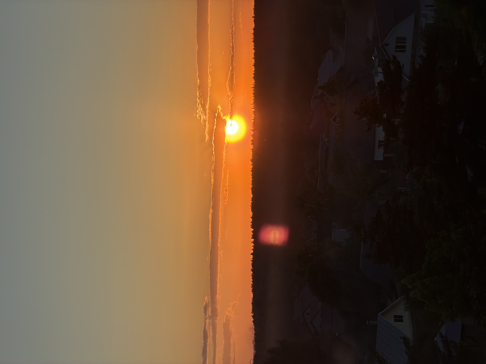 The image depicts a vibrant sunset over a landscape of houses and trees, possibly on the outskirts of an urban area. The sun is positioned near the horizon, partially obscured by a line of trees, casting a warm, orange glow across the sky. Clouds are streaked across the sky, catching the sunlight. To the right of the trees, houses are visible amidst the foliage, appearing shadowed and darker in contrast to the bright sunset. The overall scene conveys a serene and possibly suburban setting during the evening.