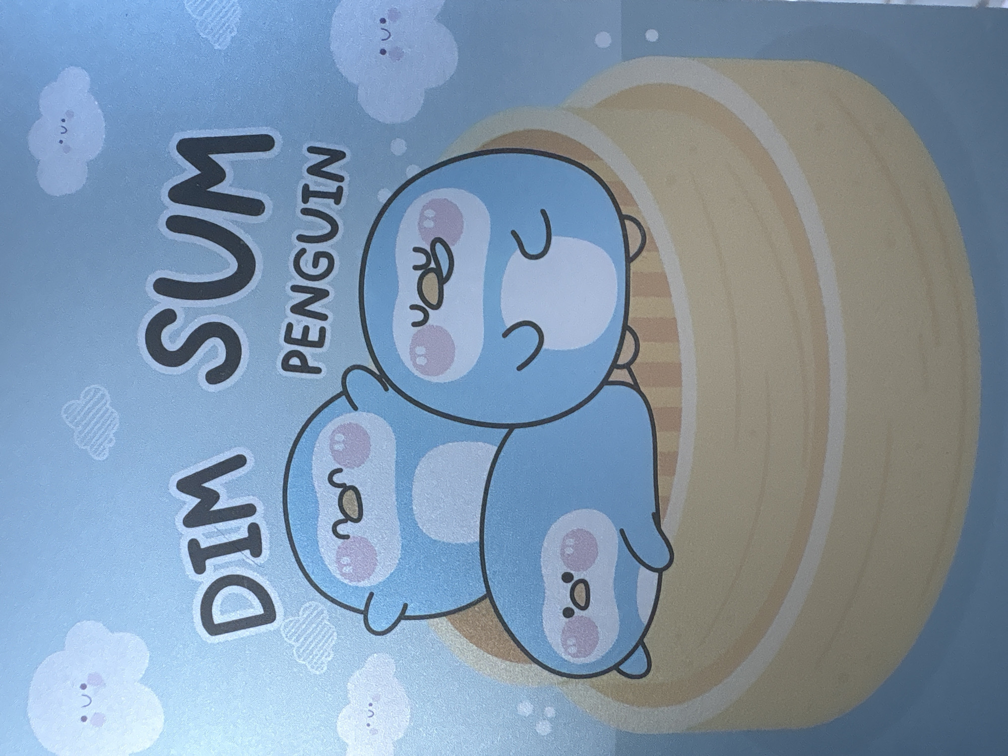 A photograph shows a light blue image with a stack of three cartoon penguins inside a tan, stacked steamer basket. The text "DIM SUM PENGUIN" is written vertically on the left side of the image in gray lettering. Small cartoon clouds are scattered throughout the blue background. The image is not of Baan Plai Laem, Thailand.