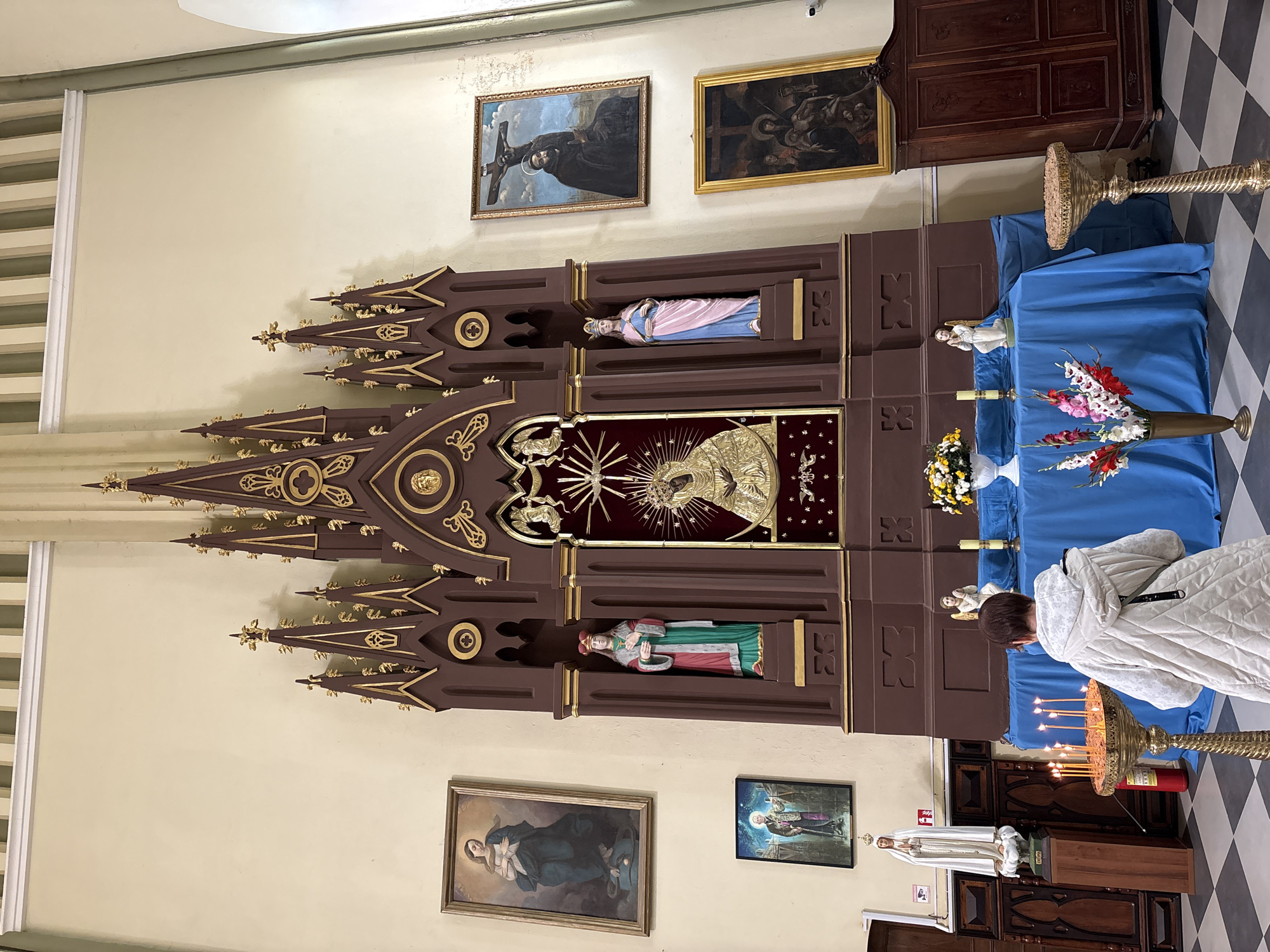 The image shows the interior of a church, likely located in Гервяцкі сельскі Савет, Belarus. In the center of the frame is a large, ornate wooden altar decorated with Gothic architectural elements and golden accents. It features several niches containing statues of religious figures. A prominent icon of the Virgin Mary, adorned with a halo and rays of light, is displayed in the central niche. To the left and right of this central icon are statues of female saints in colorful robes and headdresses. Above the central icon, a golden relief of what appears to be a cherub or angel is visible. Flanking the altar are several framed religious paintings depicting scenes from biblical narratives, including a crucifixion and a depiction of Mary with the infant Jesus. In front of the altar, a blue cloth is draped over a table adorned with floral arrangements and candles. A woman, seen from the back, is standing near a candelabra with lit candles, appearing to be in prayer or contemplation. Another statue of a robed female figure, possibly the Virgin Mary, stands to the left of the altar, near a red fire extinguisher. The floor is tiled with a black and white checkered pattern. To the right of the altar is a large, dark wooden cabinet or wardrobe. The overall ambiance is one of reverence and solemnity.