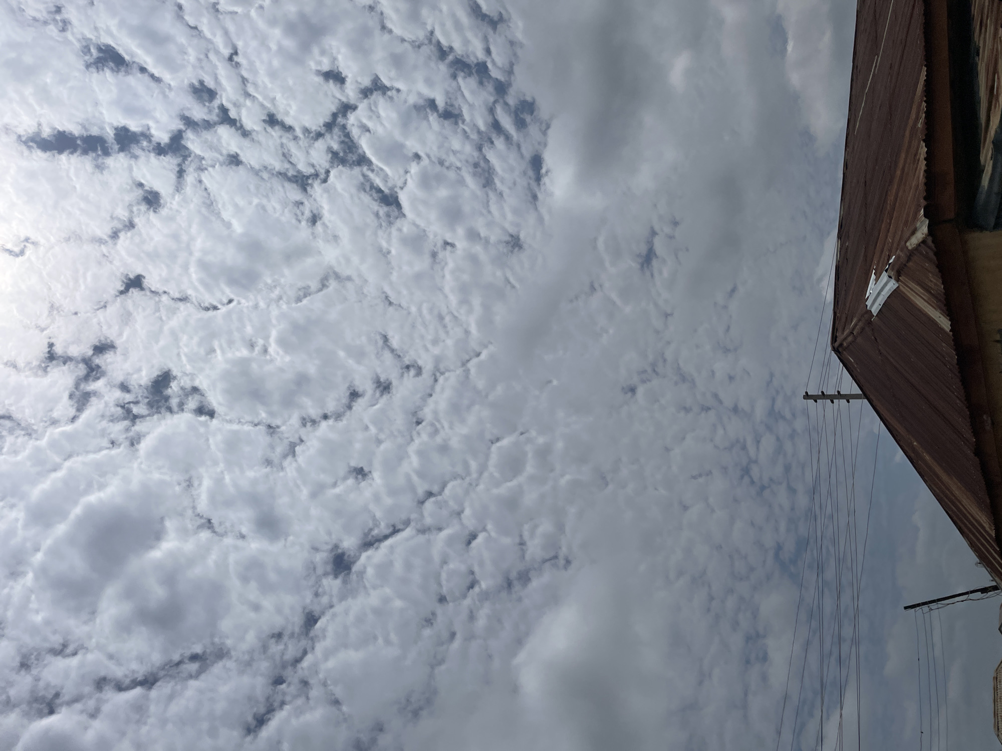 The image shows a cloudy sky above a rusted corrugated iron roof of a building in Barakin Lerawi, Nigeria. The sky is filled with scattered, puffy white clouds with patches of blue sky visible between them. Power lines are strung across the scene, with utility poles in the background. The roof of the building occupies the lower portion of the image and appears weathered and aged, with some repairs visible. The overall scene depicts an outdoor environment in what appears to be a residential or semi-urban area.