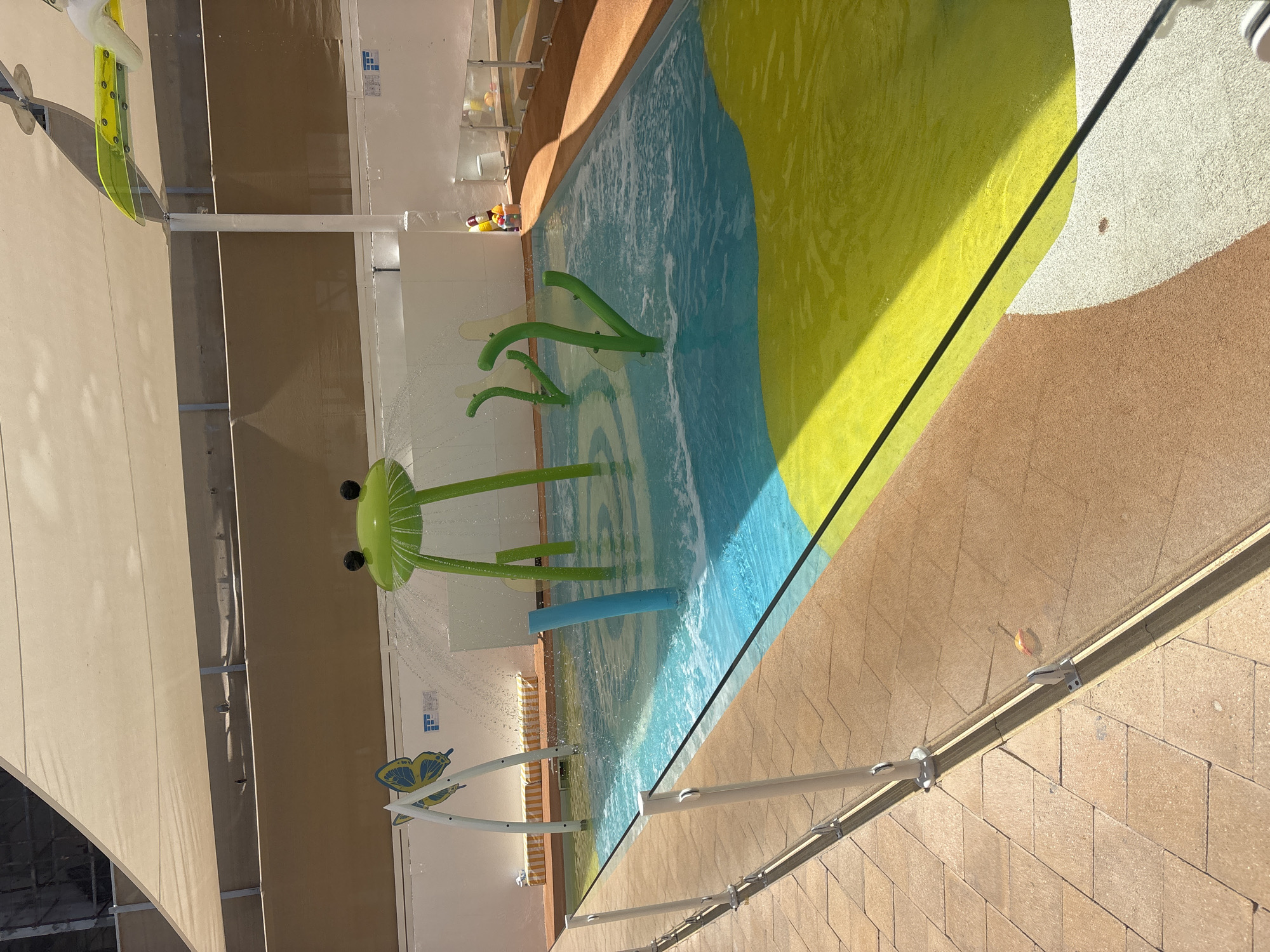 This is a photograph of a children's splash pad located in Dubai, United Arab Emirates. The splash pad features a vibrant, colorful surface with areas of blue, yellow, and white. In the water, there are several playful structures: a large green frog with water spraying from its head, and several green, plant-like sprayers. A blue pole stands in the water as well. To the left, a white arch supports a yellow and blue butterfly decoration. A clear glass fence with metal posts separates the splash pad area from a tiled walkway. The area is partially covered by a tan awning, casting shadows. The background shows a white wall and some indistinct structures. There are no people visible in the image.