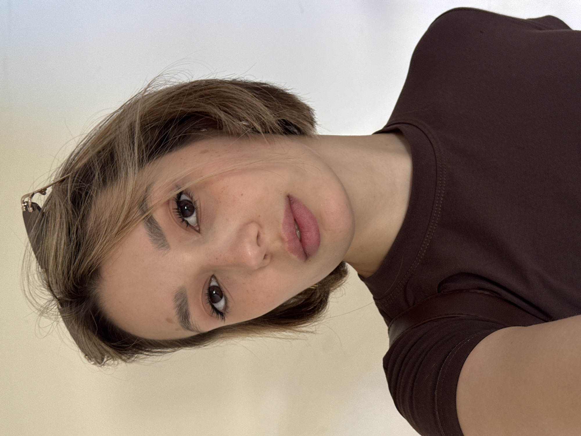 A young woman with short, blonde-streaked brown hair is looking at the camera. She has brown eyes, fair skin with freckles, and is wearing a dark brown t-shirt. Her lips are slightly parted, and she appears to be wearing makeup. There is a strap, possibly from a bag, visible over her left shoulder. The background is a plain, light-colored wall. The image is a close-up, waist-up shot.