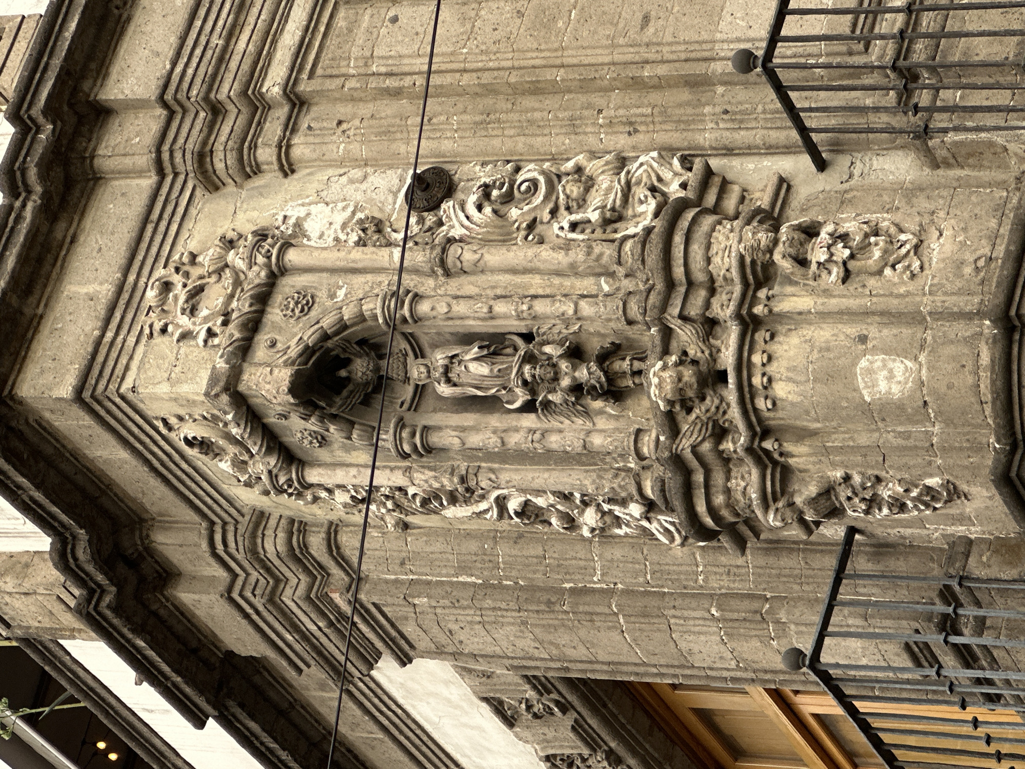 The image shows a section of highly ornate stone architecture in Mexico City, Mexico. The intricate carving features a central alcove with a figure and detailed floral motifs bordering the alcove and the surrounding structure. The building material is aged and weathered. A metal railing is visible on the upper right portion of the frame, suggesting a balcony or upper level. A window is visible in the lower right. A dark wire runs vertically through the center of the image.