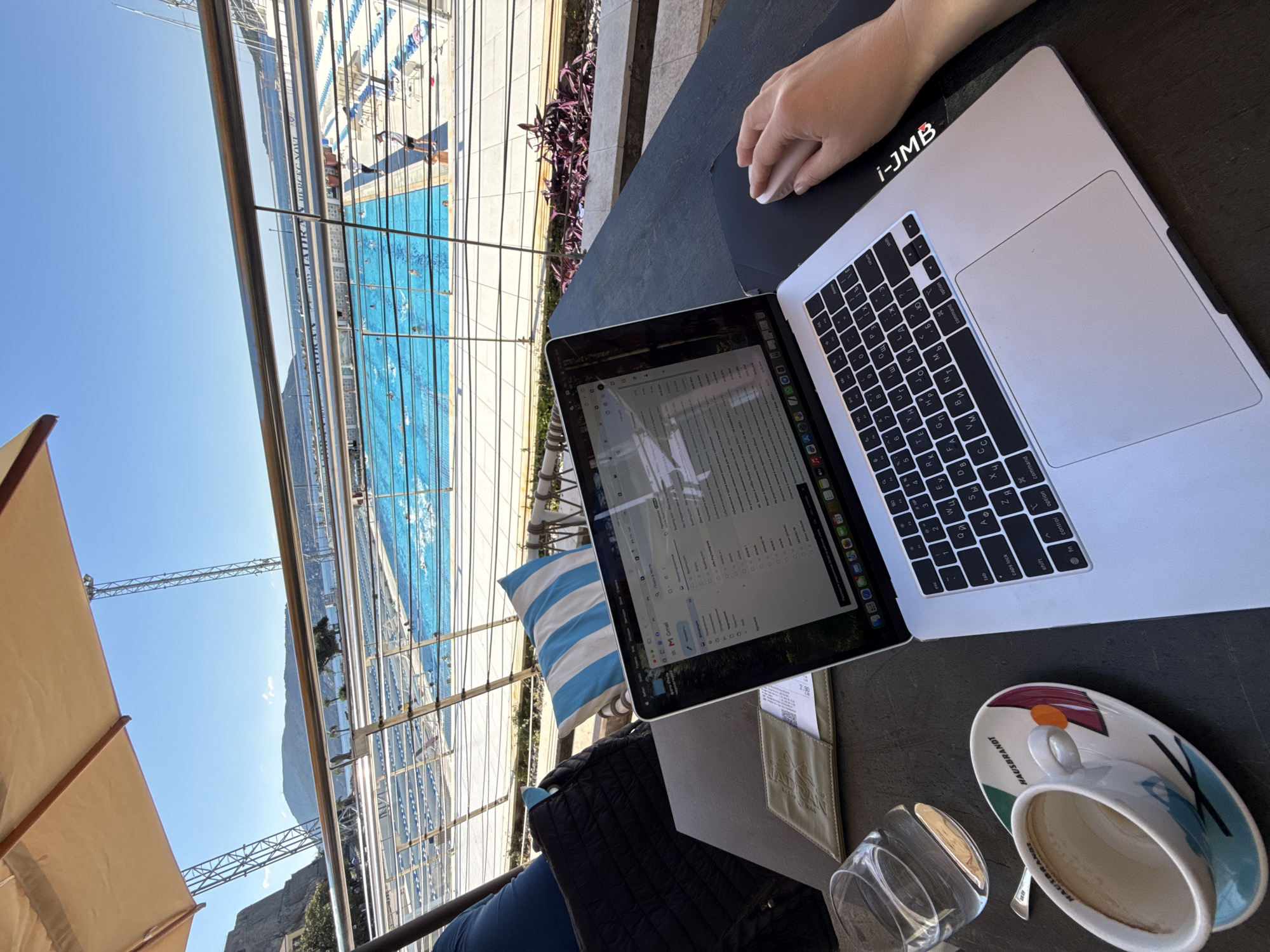 The image depicts a person working on a laptop near a swimming pool in Herceg Novi, Montenegro. The laptop screen displays what appears to be a spreadsheet or document, and a hand rests on a mouse or trackpad. The laptop is labeled with "i-JMB". A cup of coffee and a glass of water are placed on the table next to the laptop.
The swimming pool, located outside and adjacent to the seating area, appears to be of Olympic size and is surrounded by a metallic structure. People are swimming in the pool. In the background, a mountain is visible under a clear blue sky.