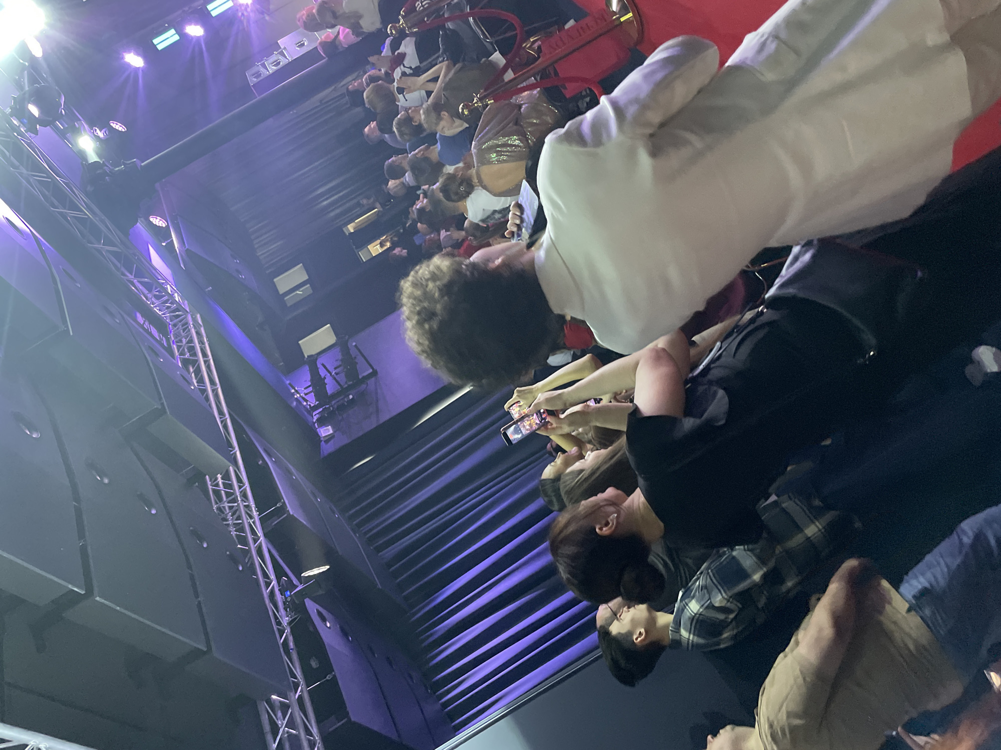 The image depicts a crowd of people at an event, possibly a premiere or launch in the city of Moscow, Russia. Many are holding up phones to take photos or videos of individuals walking along a red carpet lined with velvet ropes. These individuals appear to be celebrities or other public figures, as evidenced by the crowd's attention. The background includes stage lighting, sound equipment, and the architecture of an event space.