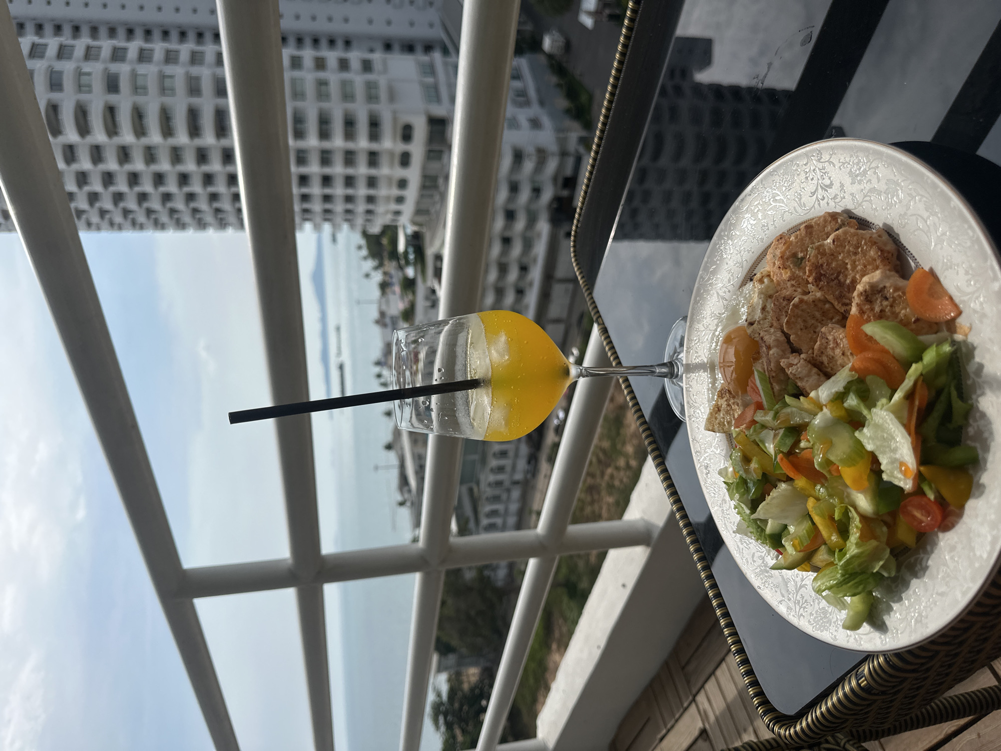 The image shows a view from a balcony in Pattaya City, Thailand, with a focus on food and drink arranged on a small table. In the foreground, there is a plate of food including what appears to be breaded cutlets, sliced carrots, and a mixed green salad. Next to the plate is a wine glass filled with an orange-colored beverage and a black straw. The balcony railing is white. In the background, there is a high-rise building and a view of the sea under a cloudy sky.
