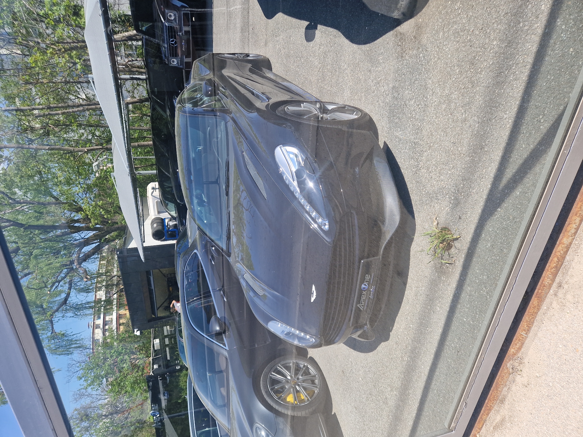 The image captures a scene through a large window or glass partition, featuring several luxury cars on a sunny day. The primary subject is a dark-colored Aston Martin sports car, likely a DBS or similar model, positioned in the foreground. Its distinctive grille, headlights with LED daytime running lights, and Aston Martin wing badge are clearly visible. On its front bumper, "APEX One" is inscribed, with smaller text underneath.
Behind the Aston Martin, reflected in the glass, is a dark Mercedes-Benz G-Class SUV. This vehicle also displays "Apex One" on its front license plate holder. Another car, possibly a Porsche due to its multi-spoke wheels and yellow brake calipers, is partially visible in the reflection on the left side of the frame.
The setting appears to be an outdoor parking area or a lot adjacent to a building, possibly a car dealership or showroom. Lush green trees are visible in the background, along with a light-colored building reflected in the glass. A covered walkway or structure extends across the upper part of the background. The ground is asphalt or concrete, with a small patch of grass seen near the bottom right. Strong shadows indicate bright, clear weather during the daytime. The overall impression is one of luxury vehicles displayed under direct sunlight.
