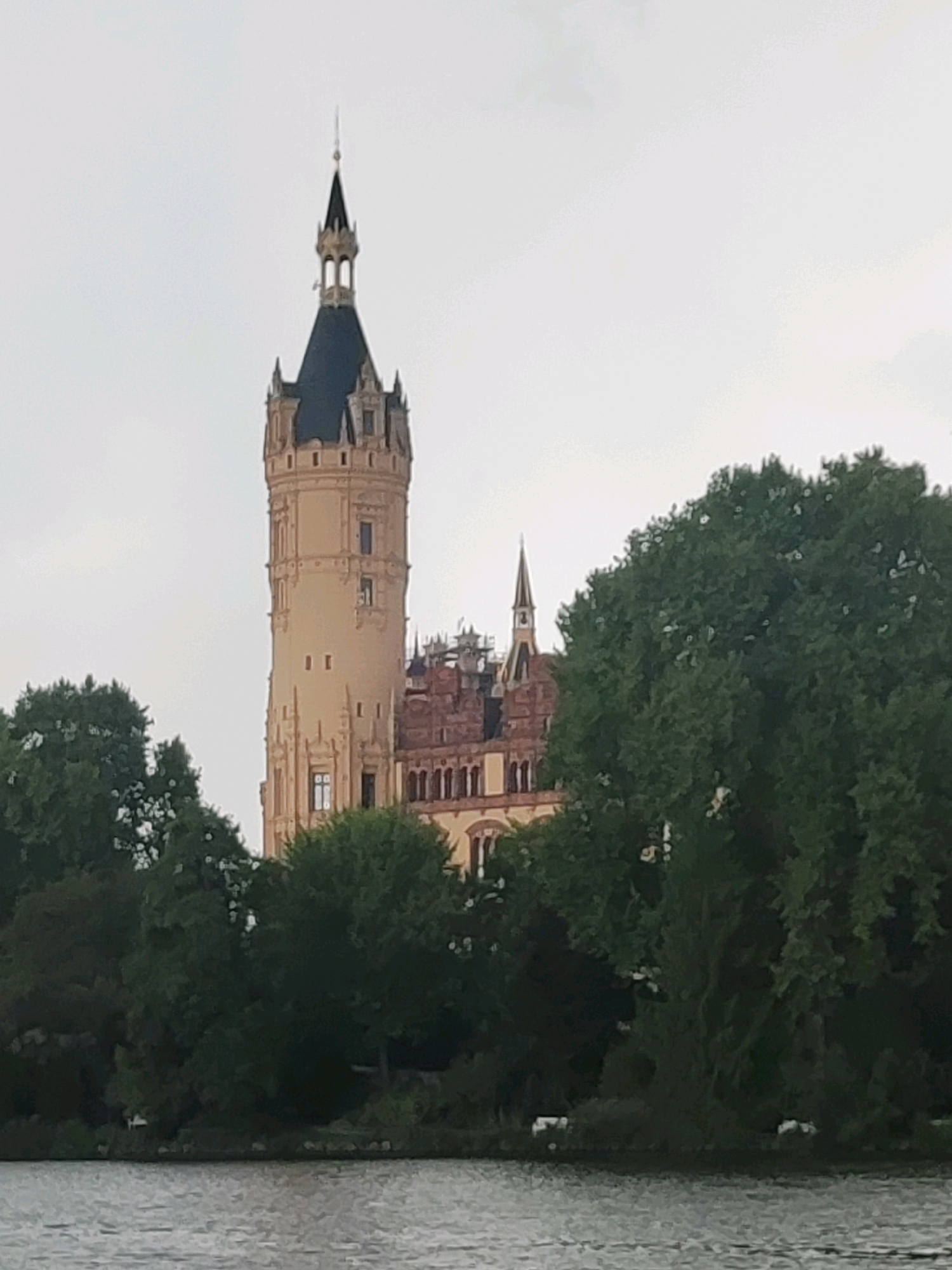 The image depicts Schwerin Castle (Schweriner Schloss), a picturesque island palace located in Schwerin, Germany. The castle, with its prominent tall tower and dark spire, is partially obscured by lush green trees in the foreground. A body of water, likely a lake, stretches across the bottom of the frame. The sky is overcast, casting a diffused light on the scene. The architectural style of the castle suggests a mix of Gothic and Renaissance influences. The composition captures the castle's imposing presence amidst a natural landscape.