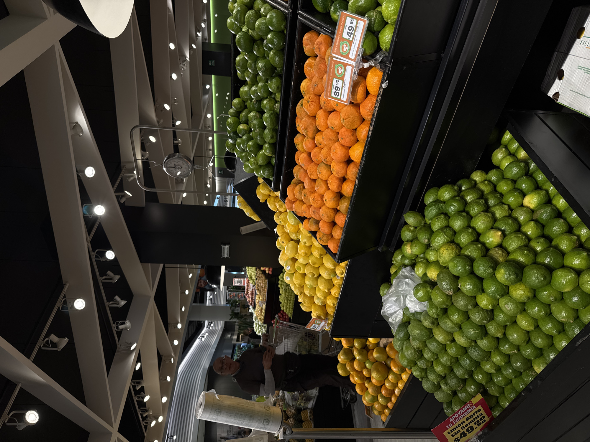 The image captures a produce section in a grocery store, likely in Puebla, Mexico, as suggested by the Spanish language signage. From the top, shelves are packed with citrus fruits: limes, oranges, and lemons. A man wearing glasses and a dark shirt is visible pushing a shopping cart. In the foreground, a roll of plastic bags sits on a metal stand. Above, the store's ceiling is characterized by a modern, geometric design of white beams and recessed lighting. Price tags are visible near the fruits, bearing Spanish text and prices.