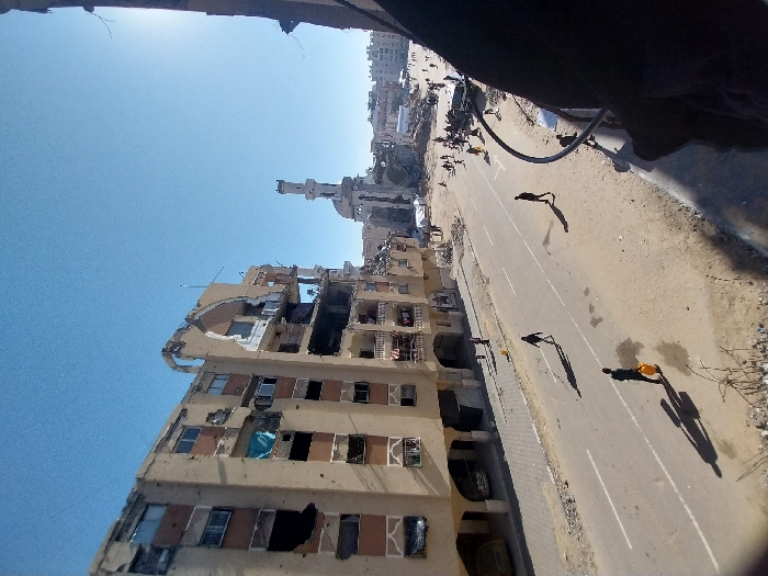 The image depicts a street scene in Khan Yunis, Palestinian Territory, under a clear blue sky. In the foreground and left, a multi-story beige residential building exhibits extensive damage, with numerous broken windows, sections of its facade missing, exposed interiors, and visible impact marks. Its upper floors, particularly an arched architectural feature, show significant structural compromise. To the right and in the mid-ground, a mosque is visible, identified by its minaret, which is visibly damaged at its apex. The mosque's main structure also appears partially destroyed.
A paved road occupies the central and lower portions of the image, featuring white lane markings and sporadic debris on its edges. Along the road and unpaved shoulders, several individuals are present. In the lower left, a person walks away from the camera, carrying a yellow jerrycan. Other individuals walk further down the street; one figure is prominent in the mid-ground, walking towards the right side of the frame. In the background, beyond the mosque, more damaged structures are discernible, and a concentration of people and debris suggests further devastation. Shadows are long, indicating either early morning or late afternoon. The perspective is elevated, looking down onto the street, with a dark structural element partially framing the lower right corner.