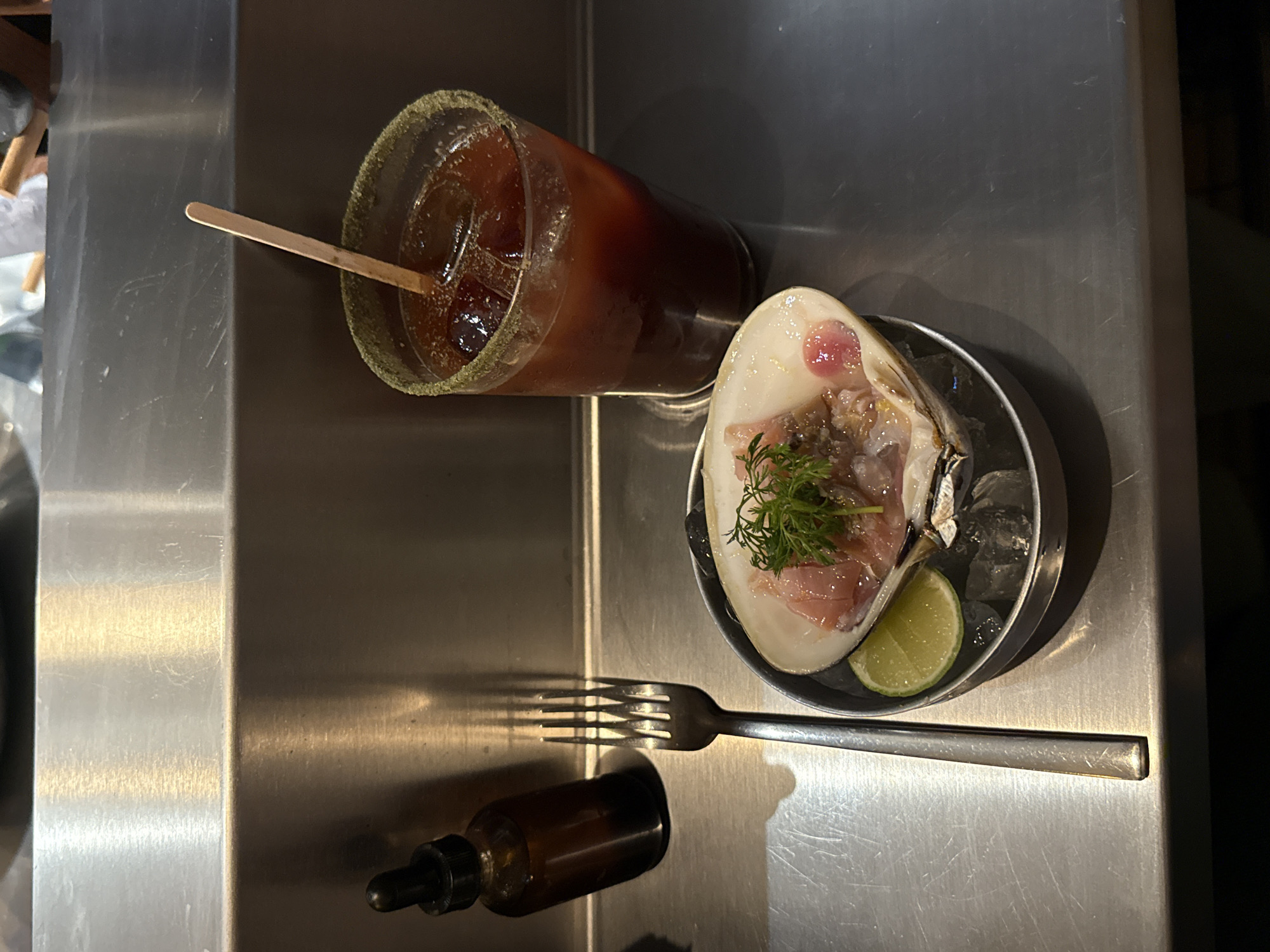 The photograph, taken from an overhead perspective, captures a culinary arrangement placed on what appears to be a stainless steel countertop, possibly within a food preparation area. A drink, likely a cocktail, resides in a glass rimmed with a green substance, and is accompanied by a wooden stir stick. Adjacent to the drink is a dish containing a large clam or oyster served over ice with a slice of lime, garnished with a sprig of dill, and possibly thinly sliced prosciutto. A fork lies nearby, along with a small bottle that may contain a sauce or seasoning. Given the subject matter, the photograph was likely taken in a restaurant or bar in Mexico City, Mexico. The specific location within the city cannot be determined solely from the image. No individuals are visible in the frame.