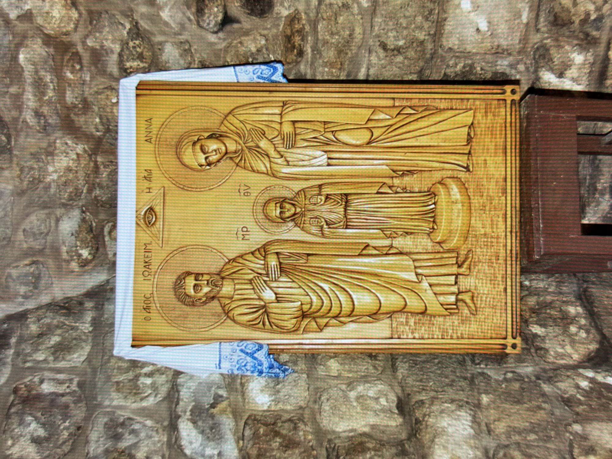 The image depicts a wooden icon of Saints Joachim and Anna, the maternal grandparents of Jesus, and their daughter Mary. The icon is carved in a traditional Byzantine style and features halos around the heads of the figures, indicating their saintly status. Saint Joachim is on the left, Saint Anna is on the right, and the young Mary is in the center. Above the figures, there is an eye within a triangle, representing the Eye of Providence. The icon is draped with a white cloth with blue embroidery, and it is mounted on a stone wall. The background appears to be an interior stone wall, possibly in a church or a similar religious setting. The icon is resting on a dark brown wooden stand. The overall scene suggests a religious context, likely within Ukraine, given the historical significance of such icons in Orthodox Christianity.