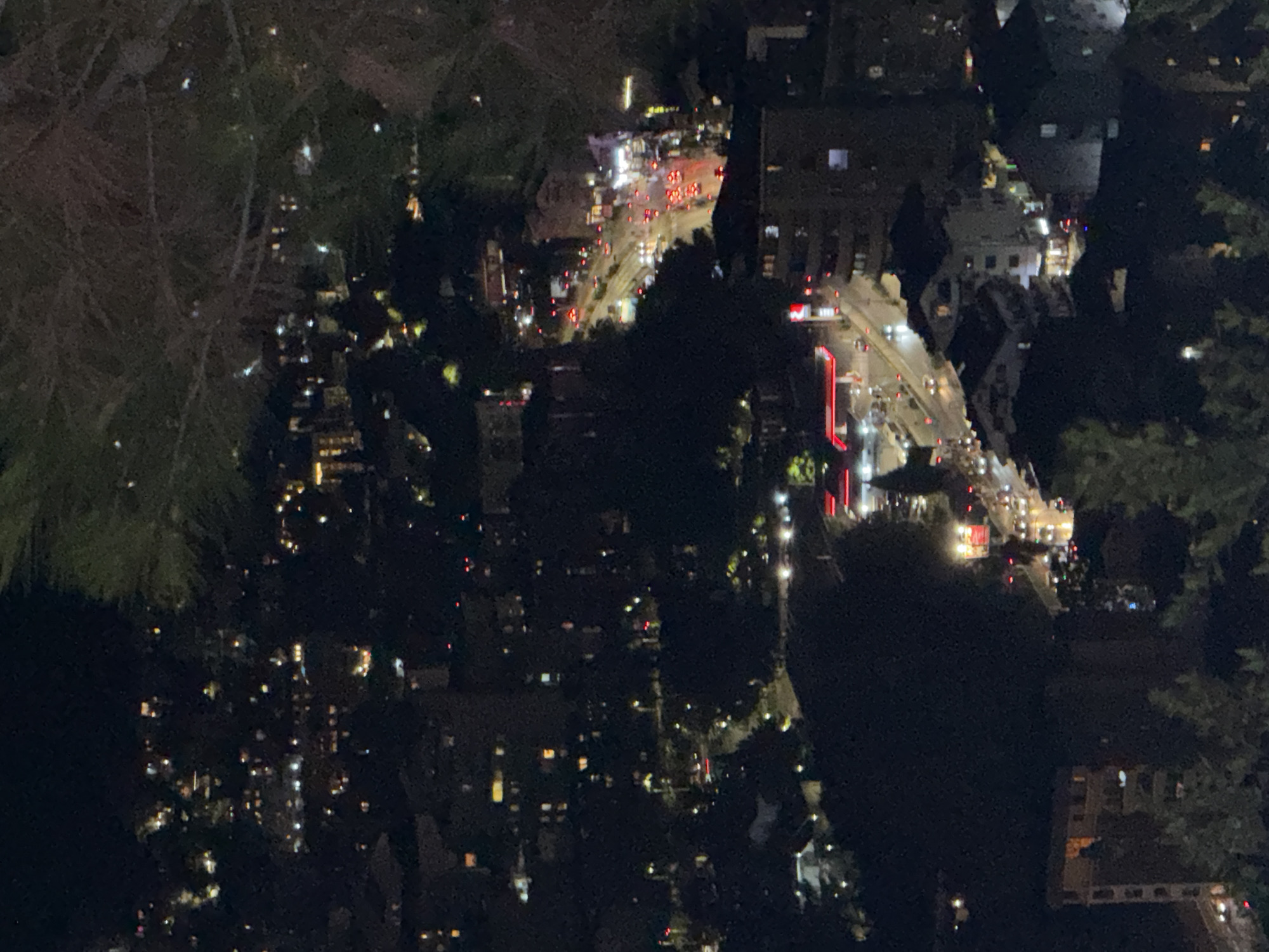 The image depicts an aerial view of a town at night. The town is characterized by numerous buildings with illuminated windows, indicating residential or commercial activity. Roads are visible, with headlights and taillights of vehicles suggesting traffic flow. A gas station with bright red awnings is a prominent feature. Trees and vegetation are interspersed throughout the urban landscape. The overall scene is one of a softly lit town under the cover of darkness. Based on the context provided, this scene is located in the city of Košljun, Montenegro.