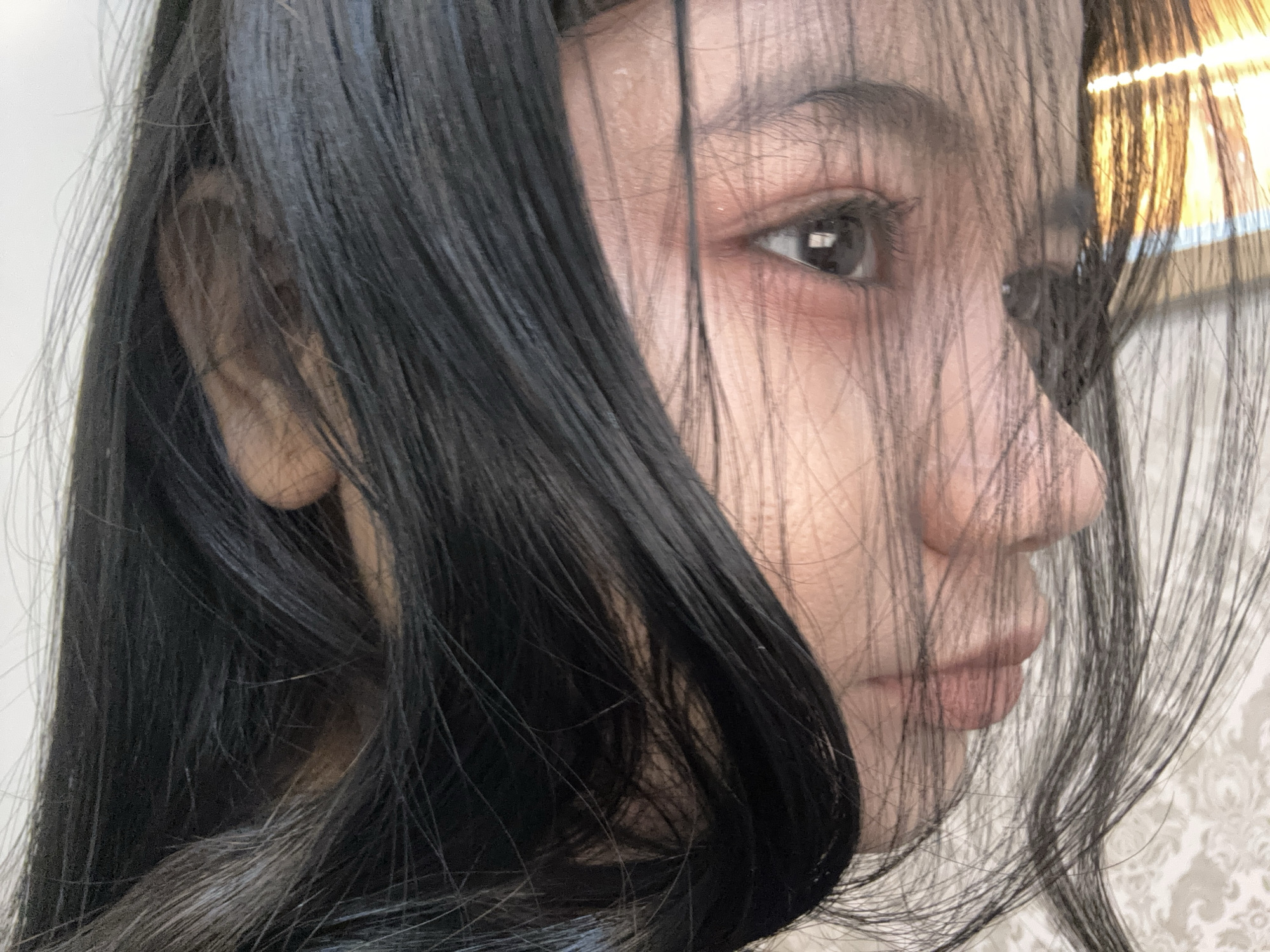 The image is a close-up, side-profile shot of a person with dark hair partially obscuring their face. The hair is long and straight, with some strands falling across the face. The visible eye is dark and almond-shaped. The person has fair skin, a slightly upturned nose, and full lips. The background is blurred, suggesting a shallow depth of field. The location information (North Jakarta, Indonesia) is not discernible from the visual content.