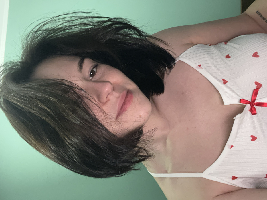 A woman with dark, short hair poses at an angle wearing a white camisole top adorned with red hearts and a red bow. Her skin appears light, and she looks directly at the camera. The background is a solid light green color. A small tattoo is visible on her upper arm. The focus is on her face and upper body.