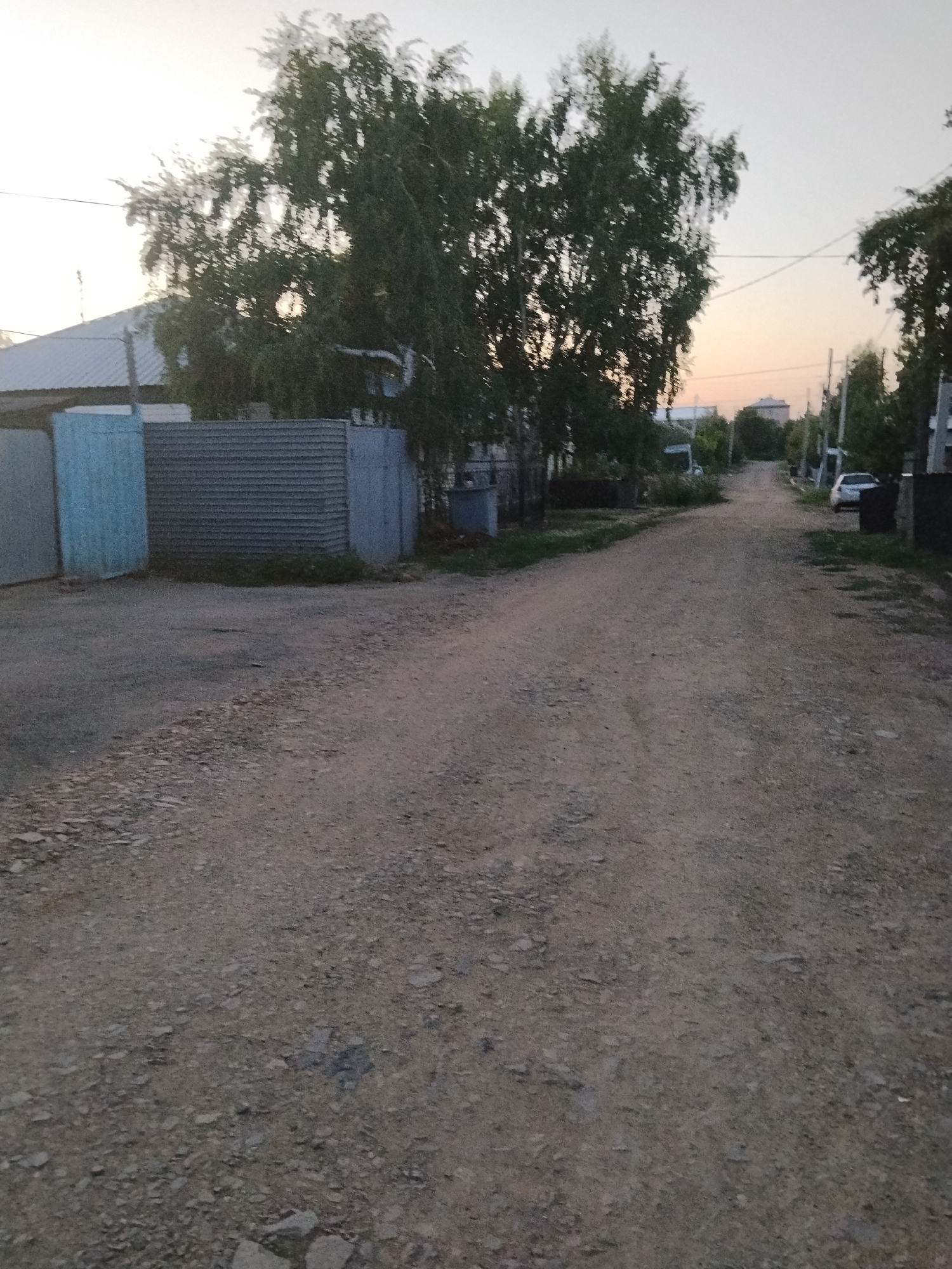A dirt road stretches into the distance, lined with trees and fences on either side. A white car is parked on the right side of the road. The sky is a pale orange, suggesting either sunrise or sunset. In the foreground, the road is covered with gravel and dirt. To the left, a grey corrugated metal fence and a light blue fence are visible. On the right, trees and houses line the road. The scene appears to be in a residential area. The image was taken in Kokshetau, Kazakhstan.
